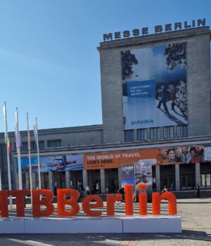 Grote rode letters "#ITBBerlin" staan voor de Messe Berlin, een congrescentrum dat rijk is aan geschiedenis, met een spandoek met de tekst "DE REISWERELD LEFT HIER". Kleurrijke vlaggen omzomen de ingang en daarboven hangt een grote toeristische poster van Alaska. | Echt Ierland Vakanties