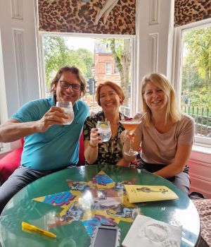 Drie mensen zitten rond een groene tafel met drankjes, lachend naar de camera. Op de tafel ligt een smartphone en een geel boek. Achter hen onthullen hoge ramen bomen en een bakstenen huis, wat doet denken aan hun onvergetelijke rondreis Ierland. Luipaardprintstof en klassieke kunstwerken sieren de muren. | Echt Ierland Vakanties