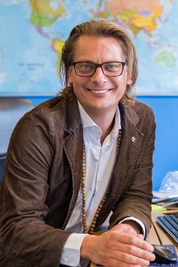 Een glimlachende persoon met een bril en schouderlang haar, gekleed in een bruin suède jasje en lichtblauw shirt, houdt een smartphone vast. Een kleurrijke kaart van Ierland is zichtbaar op de achtergrond, wat wijst op hun aankomende "rondreis Ierland", met een deel van een bureau en papieren gedeeltelijk zichtbaar. | Echt Ierland Vakanties