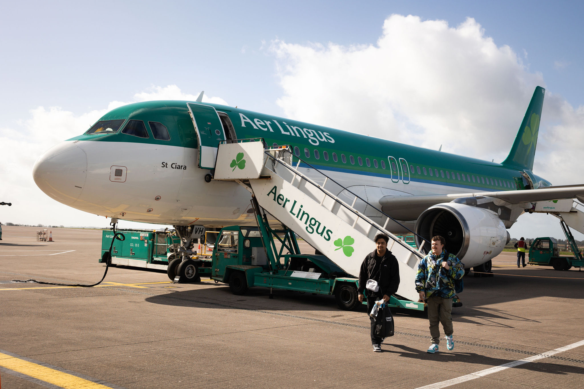 Een Aer Lingus-vliegtuig, met het opschrift "St. Ciara," staat geparkeerd op een vliegveld. Twee mensen dalen een mobiele trap af, versierd met Aer Lingus en het klavertjeslogo, en kijken uit naar hun vakantie in Ierland. Het groen-witte vliegtuig staat tegen een gedeeltelijk bewolkte lucht met bagagekarretjes in de buurt. | Echt Ierland Vakanties