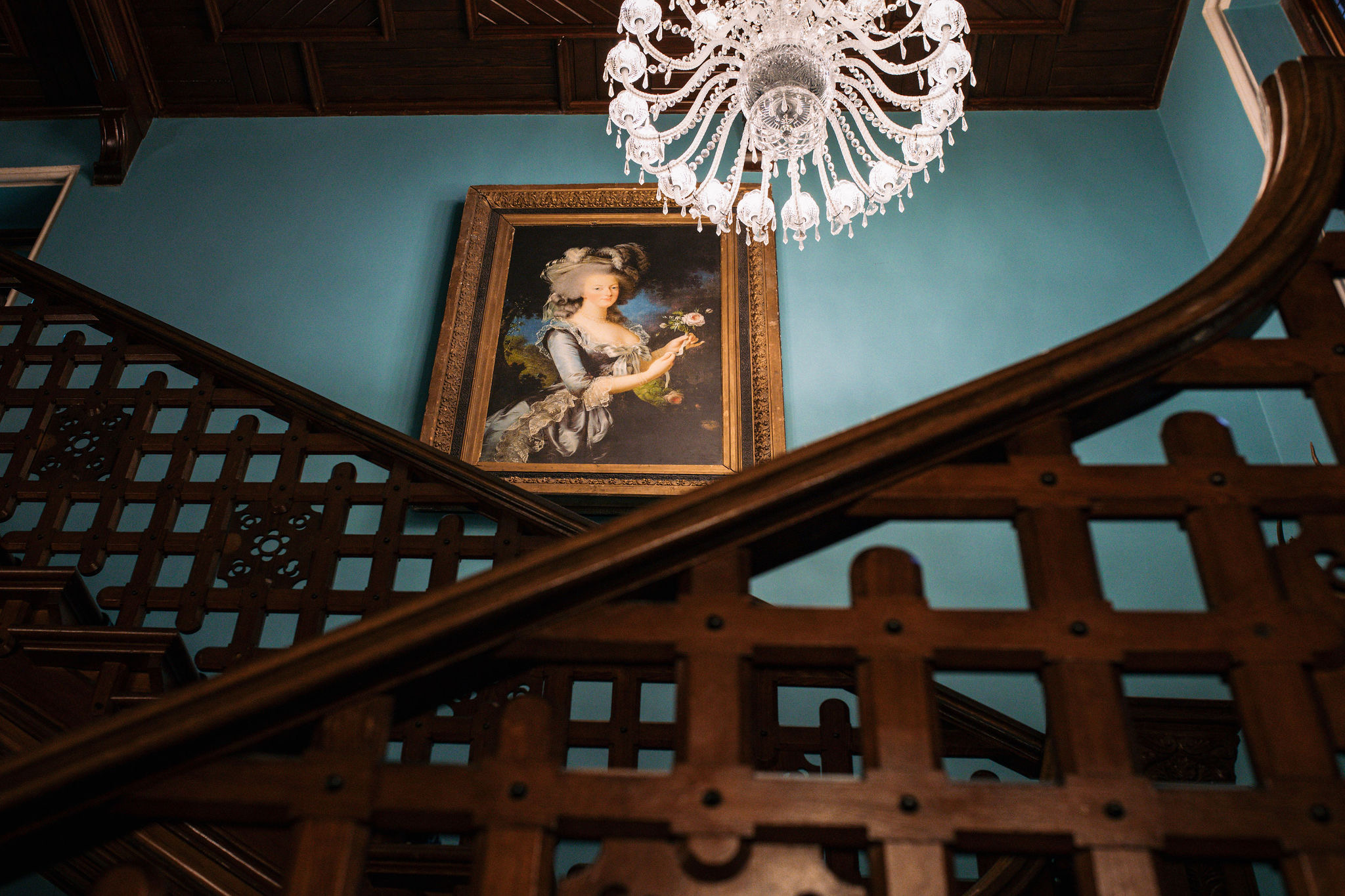 Een decoratieve houten trap met een sierlijke leuning loopt omhoog tegen een blauwgroene muur, wat doet denken aan grote huizen die je op een rondreis door Ierland zou kunnen zien. Een groot, vintage-stijl portret van een vrouw die bloemen vasthoudt hangt prominent, terwijl een elegante kristallen kroonluchter verfijning toevoegt aan de scène. | Echt Ierland Vakanties