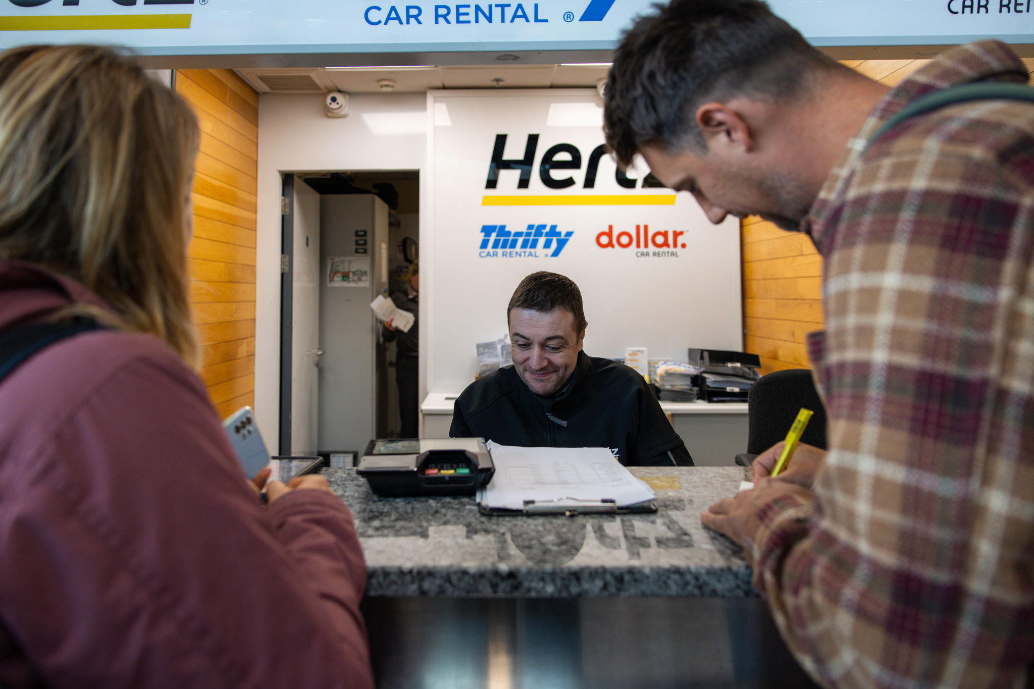 Een autoverhuurbalie met een glimlachende bediende achter een bureau helpt twee klanten bij het plannen van hun vakantie in Ierland. Het oppervlak bevat papierwerk, een rekenmachine en een pen. Borden van Hertz, Thrifty en Dollar zijn erboven zichtbaar. De klanten staan op een vloer met een patroon, met een telefoon en een pen in casual jasjes. | Echt Ierland Vakanties