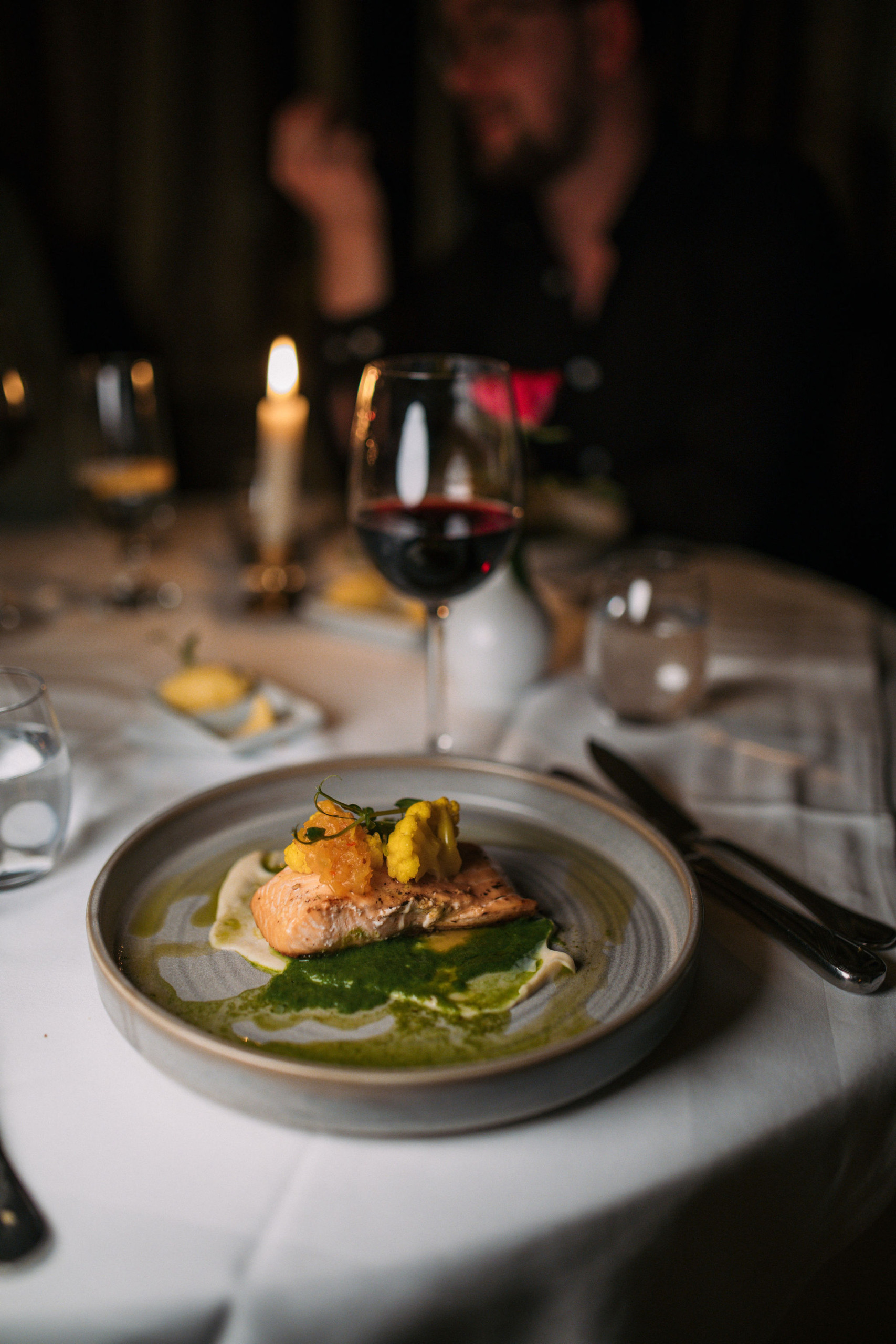 Een gastronomisch gerecht met gegrilde vis, gegarneerd met kleurrijke groenten, wordt elegant gepresenteerd tegen een ambiance die doet denken aan een rondreis Ierland. Het bord glinstert met levendige groene saus, terwijl een glas rode wijn en een brandende kaars bijdragen aan de intieme eetsfeer. | Echt Ierland Vakanties