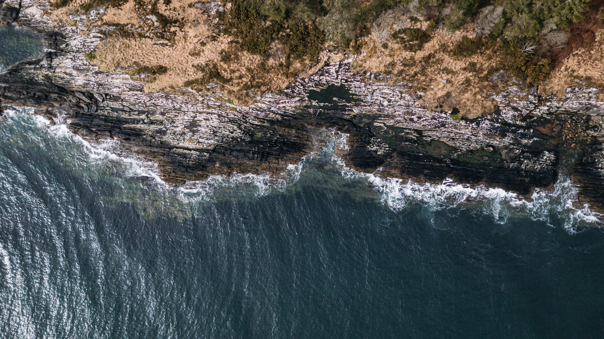 Luchtfoto van een ruige Ierse kustlijn met een donkere, golvende oceaan die grenst aan grillige, rotsachtige kliffen. De kustlijn heeft groene plekken en bruine, grasachtige gebieden. Wit zeeschuim vormt zich langs de rotsen waar de golven breken, wat adembenemende vergezichten biedt voor uw vakantie in Ierland. | Echt Ierland Vakanties
