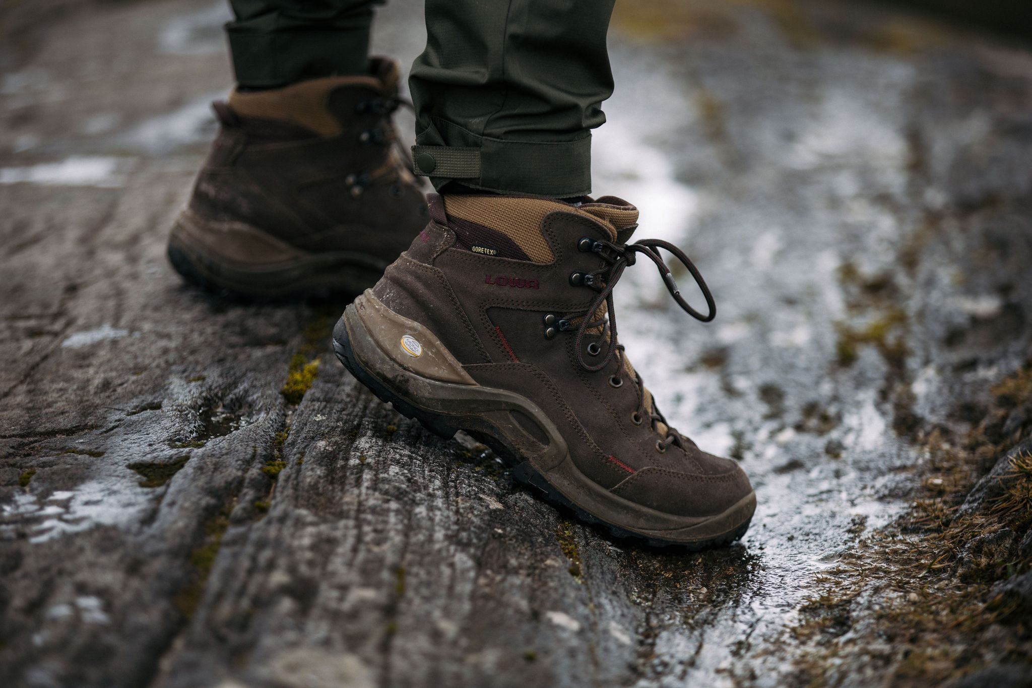 Close-up van een persoon in bruine wandelschoenen met rode accenten, staand op een natte rots. De veterlaarzen, gecombineerd met licht opgerolde groene broeken, hinten naar avonturen zoals die op vakanties in Ierland. Met mos bedekte rotsen glinsteren van het water en vangen de ruige aantrekkingskracht van een vakantie in Ierland. | Echt Ierland Vakanties