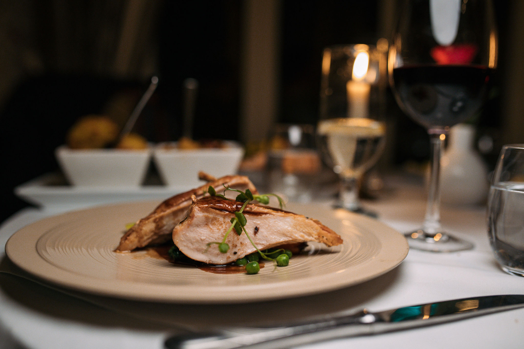 Gourmet gerecht met gesneden kipfilet gegarneerd met groene kruiden op een beige bord, wat doet denken aan smaken van een onvergetelijke vakantie in Ierland. Een glas rode wijn, een brandende kaars en een waterglas sieren de achtergrond. Zachte verlichting creëert een elegante eetsfeer, met bijgerechten zichtbaar op tafel. | Echt Ierland Vakanties