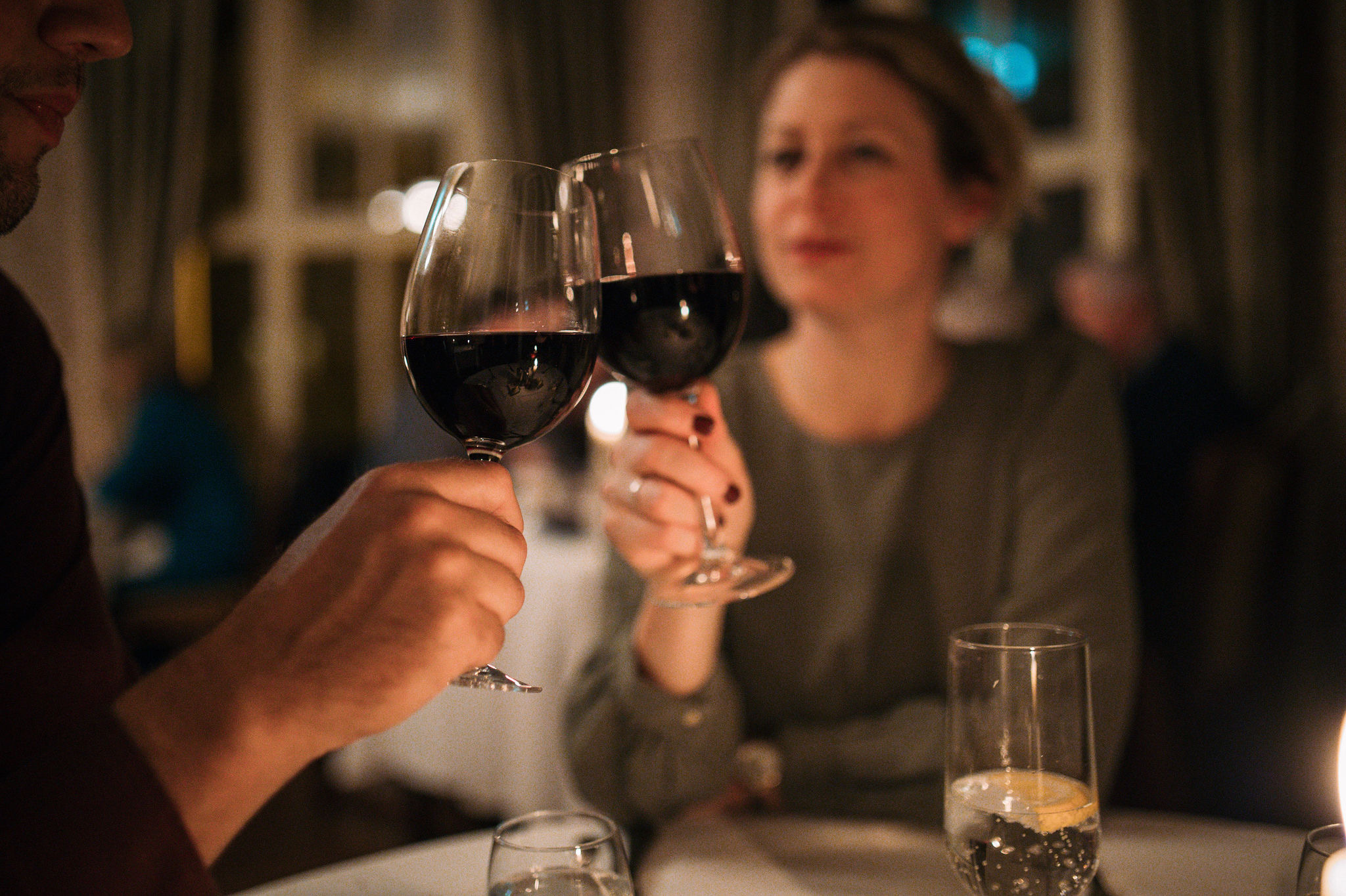 Een man en een vrouw klinken met rode wijn in een slecht verlicht restaurant, om hun recente vakantie in Ierland te vieren. Het gezicht van de vrouw is lichtjes wazig en ze draagt een groene trui die doet denken aan Ierse landschappen. Een kaars gloeit op tafel, met gordijnen die de drukke achtergrond zachtjes omlijsten. | Echt Ierland Vakanties