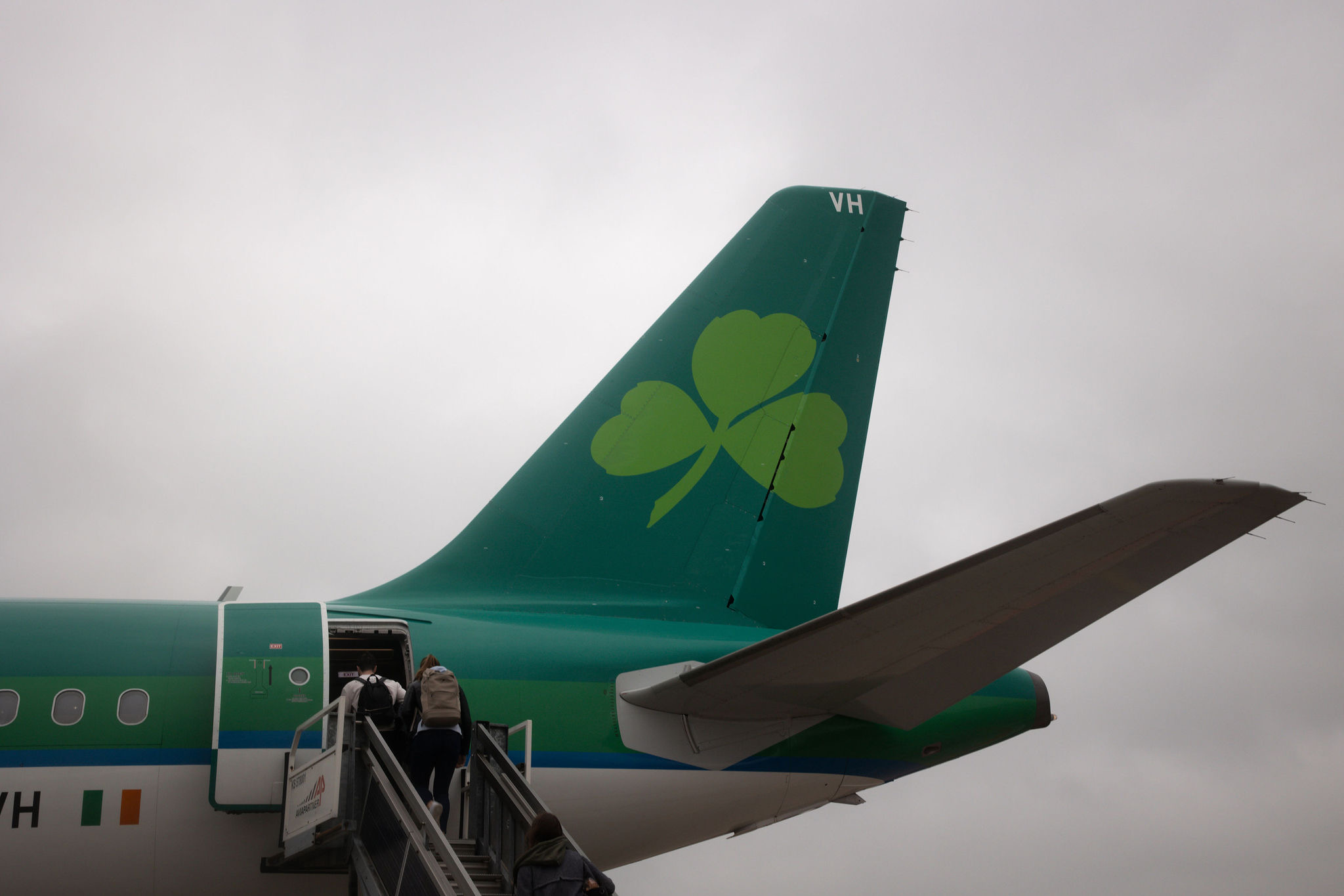 De afbeelding toont de staart van een vliegtuig met een groene klaver, wat aangeeft dat het Aer Lingus is, een ideaal beginpunt voor een rondreis door Ierland. Passagiers stappen op een bewolkte dag via een trap aan boord. De romp van het vliegtuig is groen en wit geverfd, met een kleine Ierse vlag bij de deur, perfect voor degenen die op zoek zijn naar een vakantie in Ierland. | Echt Ierland Vakanties