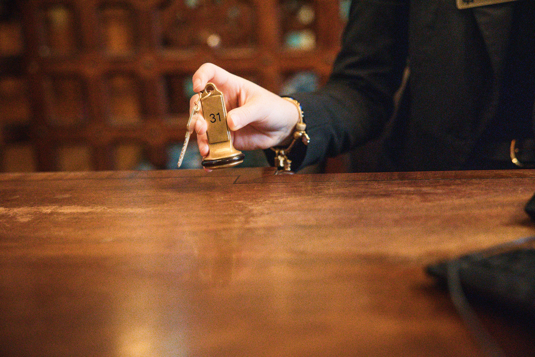 Een persoon in een donker pak strekt zijn hand uit over een houten toonbank, met een hotelkamersleutel met "31" gegraveerd op een gouden sleutelhanger. De achtergrond is voorzien van wazig hout, wat de warmte van een gezellige herberg op een rondreis Ierland weerspiegelt, wat een uitnodigende sfeer creëert. | Echt Ierland Vakanties