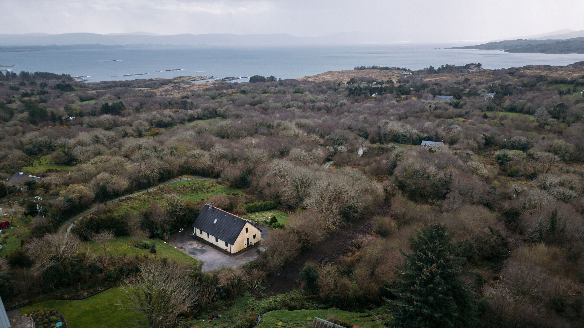 Luchtfoto van een landelijk landschap in de stijl van een serene vakantie in Ierland. Een enkel beige huis is genesteld tussen dichte, kale bomen, met een oprit en een klein gazon. In de verte zijn heuvels en een uitgestrekt waterlichaam zichtbaar onder een bewolkte lucht. | Echt Ierland Vakanties
