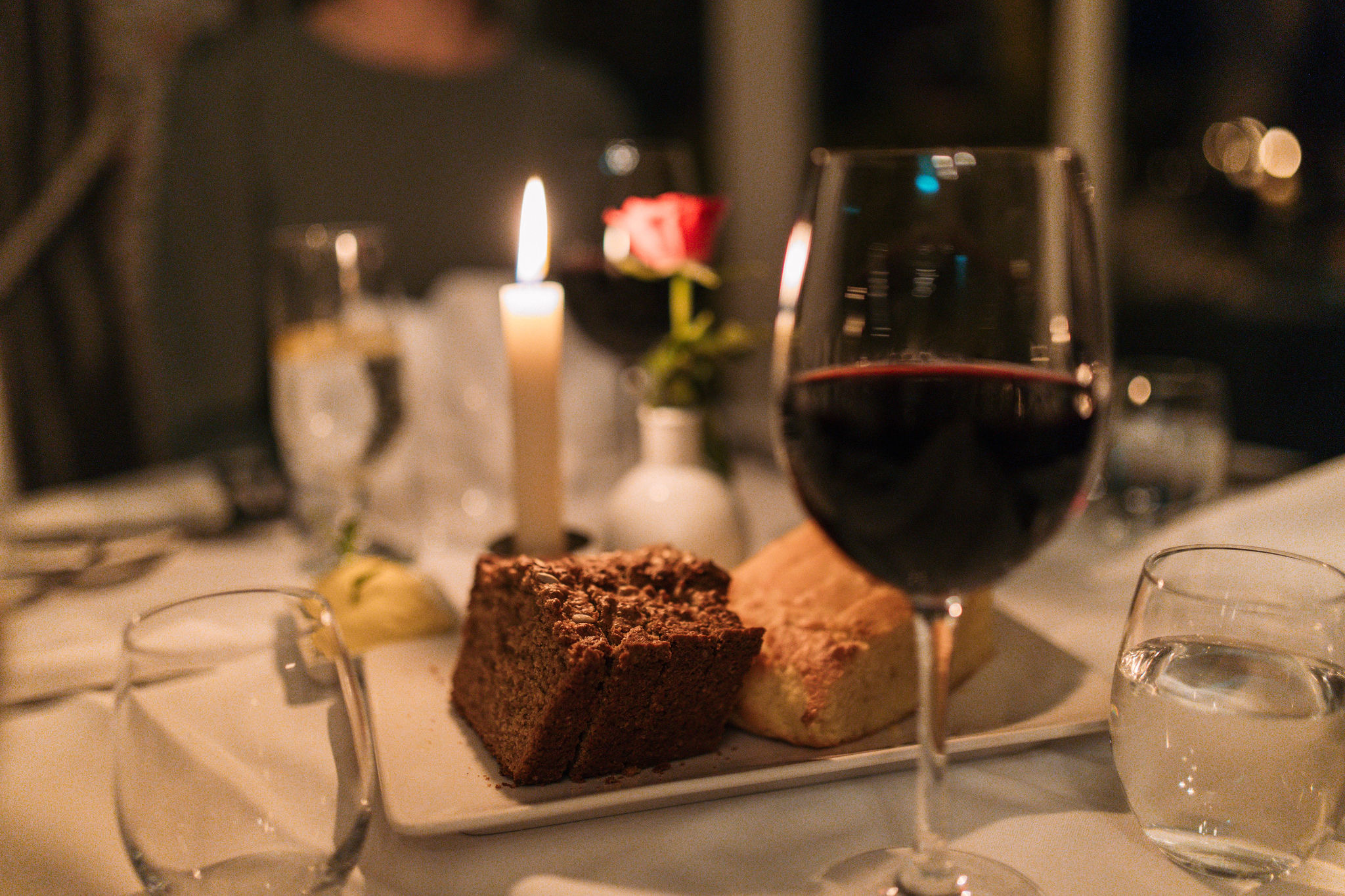 Een tafel bij kaarslicht roept de charme op van een vakantie in Ierland, met een glas rode wijn op de voorgrond. Daarachter staat een wit bord met chocoladetaart en brood. Een brandende kaars en een vaas met een rode roos zorgen voor ambiance, terwijl glazen water op het witte tafelkleed rusten. | Echt Ierland Vakanties