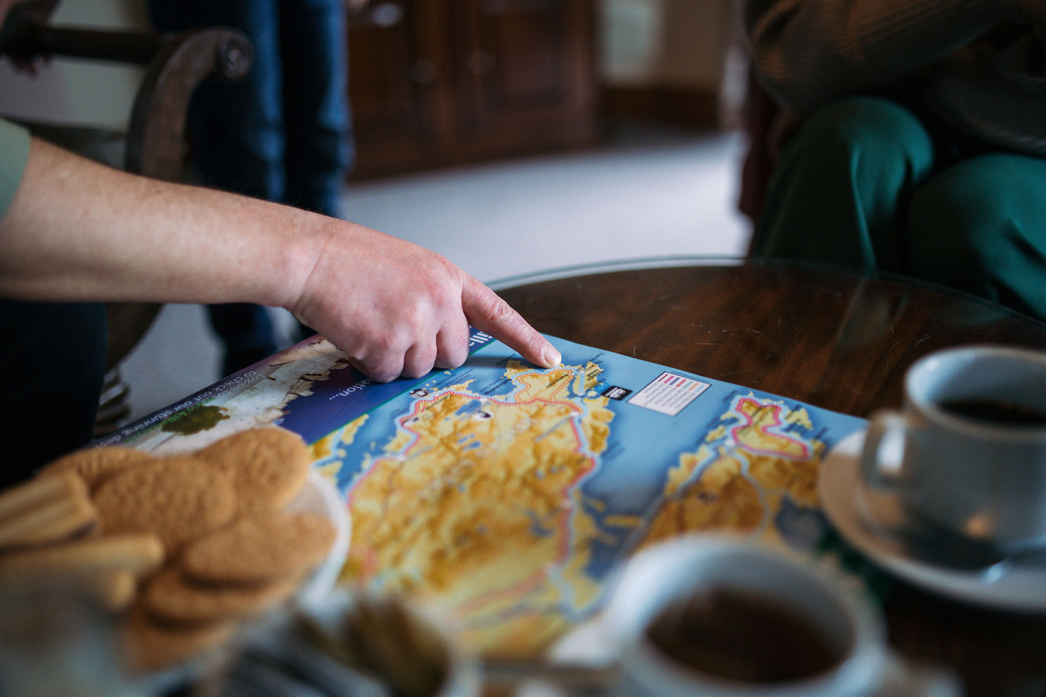 Een persoon wijst naar een locatie op een kleurrijke kaart op een glazen tafel, mogelijk zijn rondreis Ierland plannend. Naast de kaart nodigen kopjes koffie en een bord koekjes uit tot een lang gesprek. Andere mensen zitten in de buurt, omlijst door een houten kast en zacht wazig meubilair op de achtergrond. | Echt Ierland Vakanties