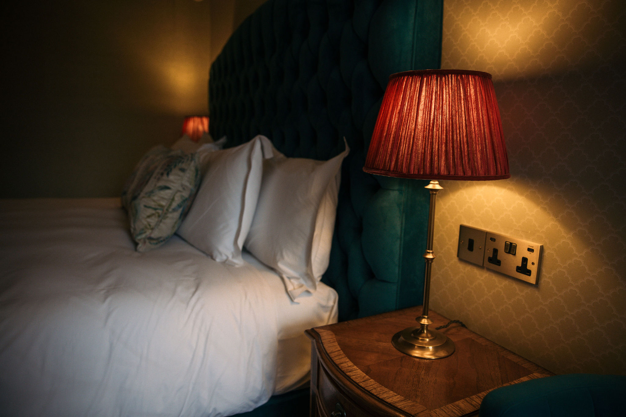 Een knusse slaapkamer, die doet denken aan een vakantie in Ierland, heeft een netjes opgemaakt bed met wit linnen en kussens met patronen tegen een gecapitonneerd groen hoofdeinde. Een houten nachtkastje heeft een gouden lamp met een rode geplooide kap, die een warme gloed werpt op de behangen muur met patroon en een stopcontact. | Echt Ierland Vakanties