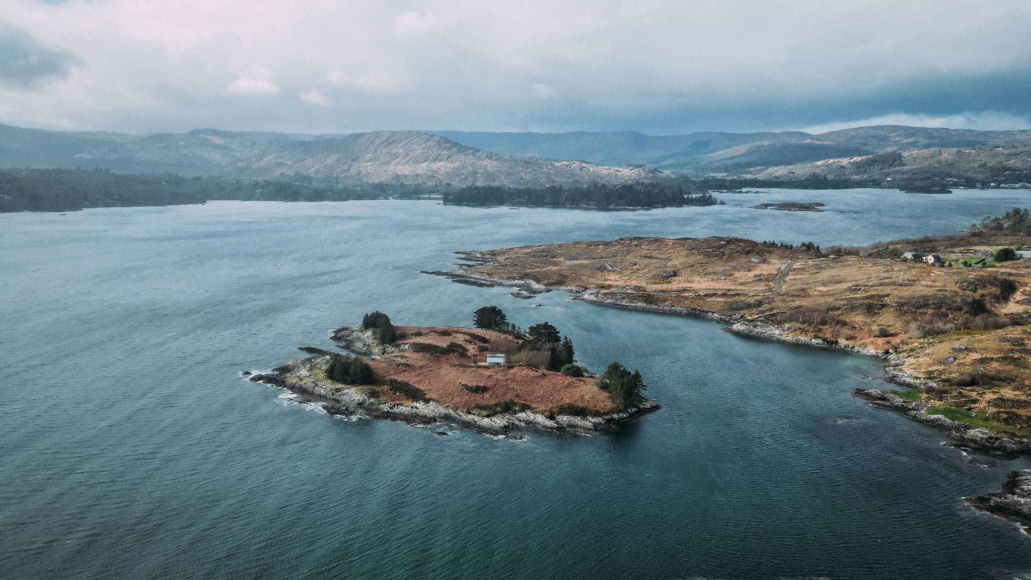 Luchtfoto van een klein, rotsachtig eiland met schaarse bomen en struiken, omgeven door een kalm waterlichaam. Heuvels en een bergachtig landschap vormen de achtergrond onder een bewolkte lucht, wat een vredig en ruig natuurlandschap creëert - een ideaal hoogtepunt voor elke rondreis Ierland. | Echt Ierland Vakanties
