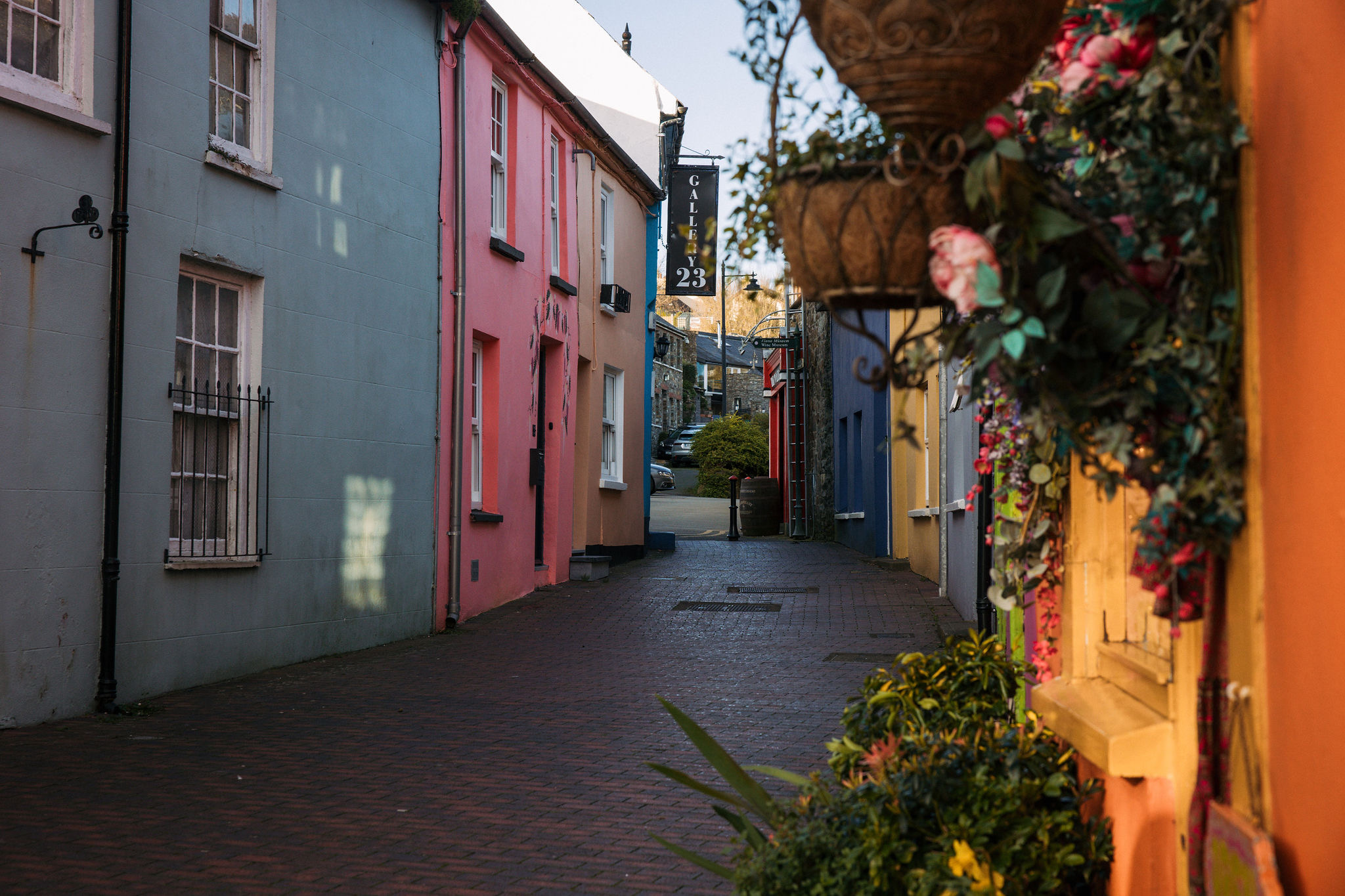 Smal steegje met kleurrijke gebouwen aan beide kanten, met pastelblauwe, roze en oranje gevels die typerend zijn voor een charmante rondreis Ierland. Hangende manden met bloemen staan aan de rechterkant. Het geplaveide pad leidt naar een klein straatje op de achtergrond, met een bord waarop "23" staat. Zacht zonlicht verlicht het tafereel. | Echt Ierland Vakanties