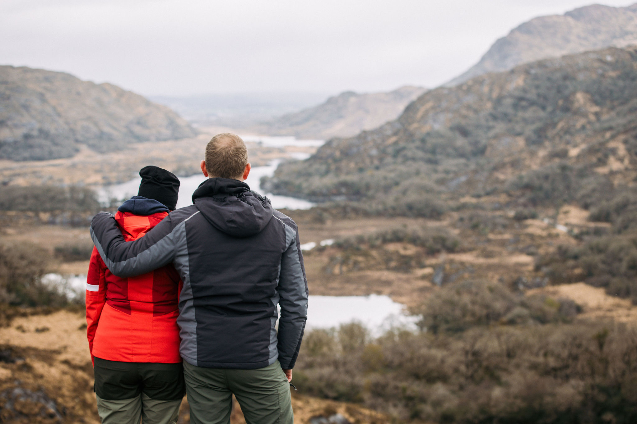 Twee mensen staan met hun rug naar de camera op een heuvel, onderdeel van een rondreis Ierland, met uitzicht op een schilderachtige vallei. De een draagt een rood jasje, de ander in het donker, met een arm om hen heen. Het landschap bestaat uit glooiende heuvels, weelderige stukken groen en een kronkelende rivier onder een bewolkte lucht. | Echt Ierland Vakanties