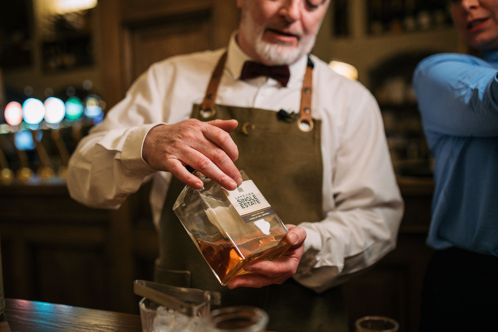 Een man met een bruin schort en een wit shirt schenkt whisky uit een fles met het label "Single Estate" terwijl hij verhalen vertelt over zijn rondreis Ierland. Op de achtergrond is een bar met glazen en taps. Hij draagt een netjes bijgesneden baard, een kastanjebruine vlinderdas en een andere figuur in het blauw is gedeeltelijk naast hem zichtbaar. | Echt Ierland Vakanties