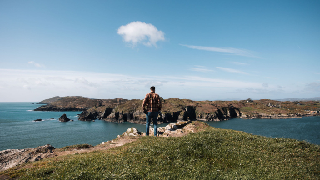 Een persoon in een geruit overhemd en jeans staat op een met gras begroeide klif, genietend van het adembenemende kustlandschap tijdens hun vakantie in Ierland. De kalme zee contrasteert met de ruige kliffen die het water in reiken, terwijl een paar wolken de helderblauwe lucht bedekken, wat dit schilderachtige uitzicht versterkt. | Echt Ierland Vakanties