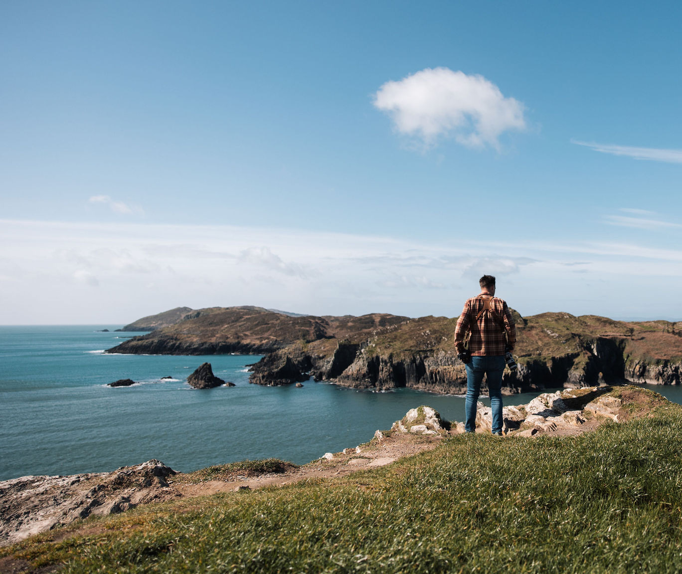 Een persoon in een geruit overhemd en jeans staat op een met gras begroeide klif, genietend van het adembenemende kustlandschap tijdens hun vakantie in Ierland. De kalme zee contrasteert met de ruige kliffen die het water in reiken, terwijl een paar wolken de helderblauwe lucht bedekken, wat dit schilderachtige uitzicht versterkt. | Echt Ierland Vakanties