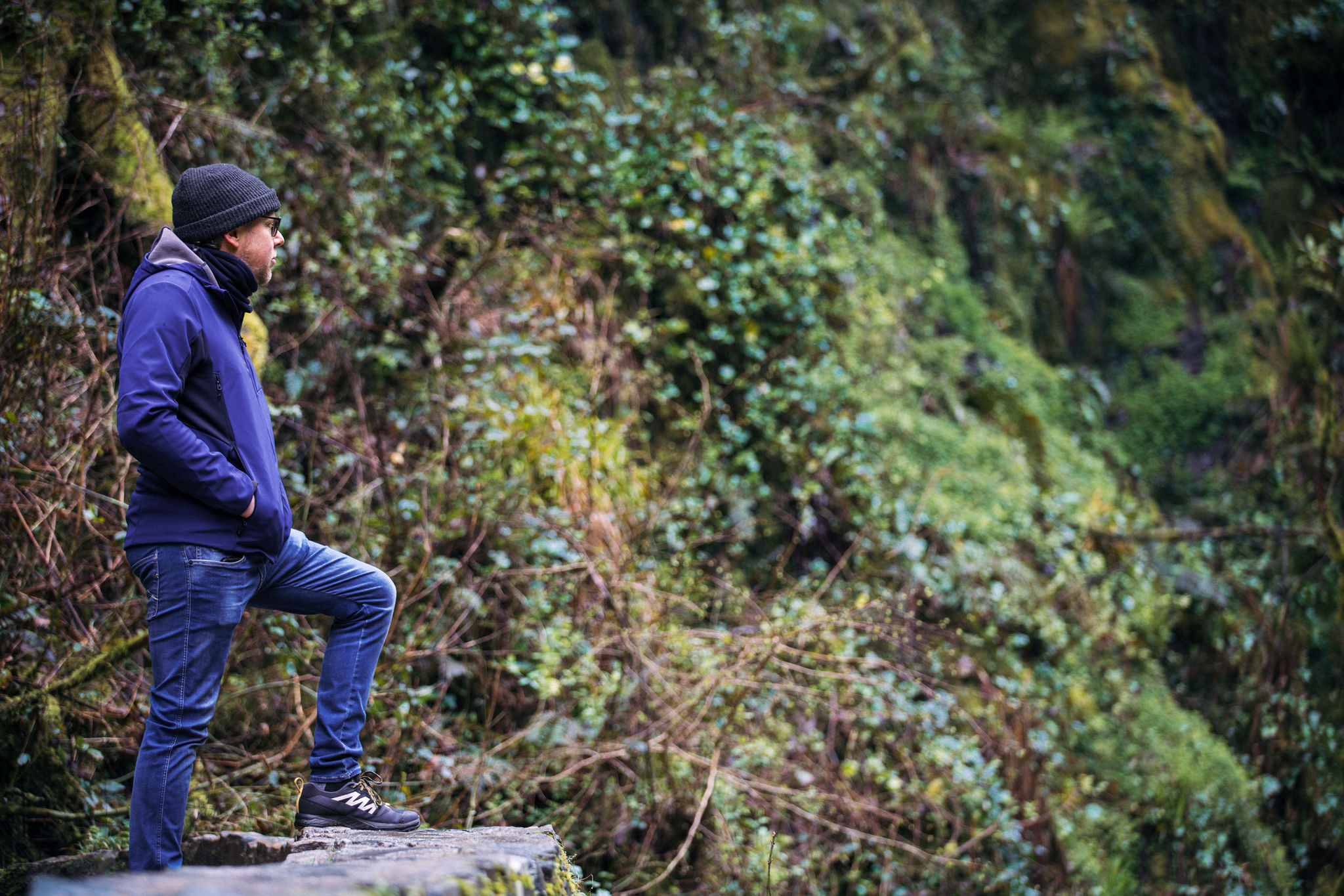 Een man staat op een rotsrichel in een bos tijdens zijn rondreis Ierland, gekleed in een donker jasje, jeans, sneakers en een muts. Hij staart in de verte, met weelderig groen gebladerte en met mos bedekte rotsen op de achtergrond, wat een serene en natuurlijke setting creëert. | Echt Ierland Vakanties