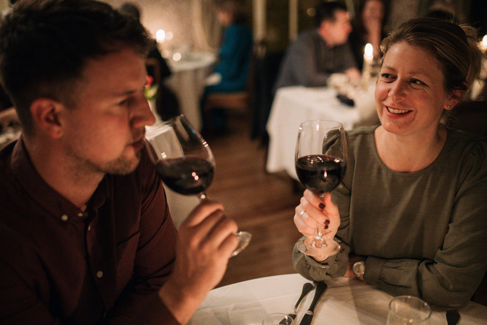 Een man en een vrouw zitten aan een eettafel in een schemerig restaurant, beiden met een glas rode wijn in de hand. De vrouw, gekleed in een groen topje dat doet denken aan een onvergetelijke vakantie in Ierland, glimlacht naar de man in een kastanjebruin shirt. Andere gasten creëren een warme, intieme ambiance in de zacht gefocuste achtergrond. | Echt Ierland Vakanties