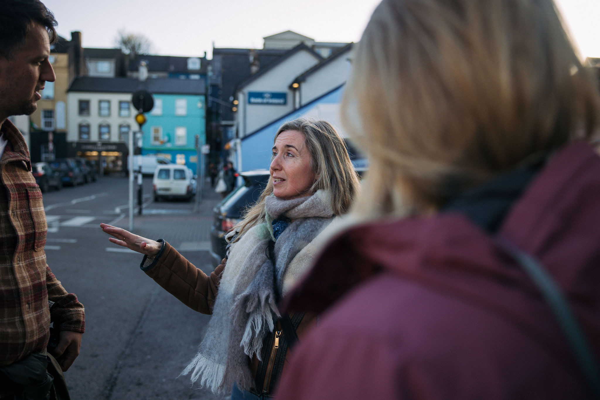 Een vrouw met lang haar en een grote sjaal gebaart geanimeerd terwijl ze haar vakantieplannen voor Ierland bespreekt met een man in een geruit overhemd en een ander persoon in een rood jasje. Ze staan op een levendige straat met kleurrijke gebouwen, die de charme belichamen die je tijdens een rondreis Ierland kunt vinden. | Echt Ierland Vakanties