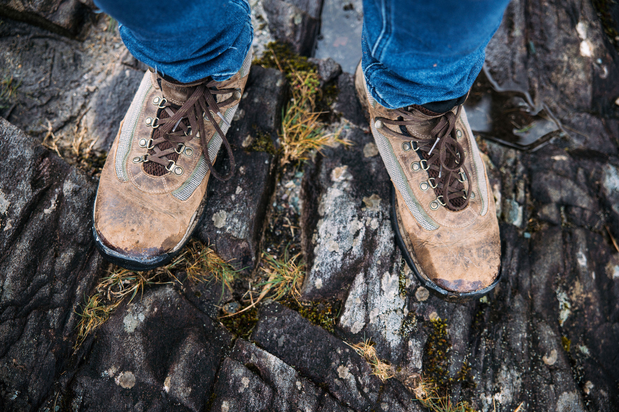 Close-up van een persoon in versleten, bruine wandelschoenen op rotsachtig terrein, de veters strak vastgebonden van een ruige rondreis Ierland. Blauwe spijkerbroeken steken uit als stukken gras en kleine plassen op de getextureerde rotsen, die de essentie van een avontuurlijke vakantie Ierland vastleggen. | Echt Ierland Vakanties