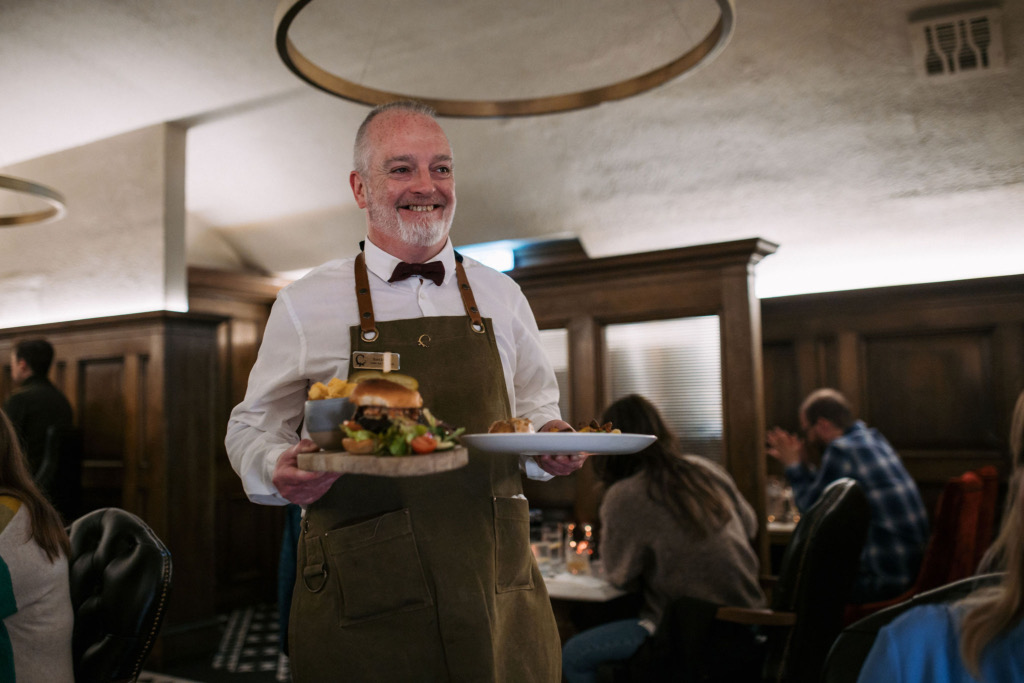 Een ober met een baard en een vlinderdas, die een olijfgroene schort draagt, glimlacht terwijl hij borden met eten vasthoudt. De gedimde verlichting en houten accenten van het restaurant roepen de warmte op die doet denken aan vakantie in Ierland, met gasten die op de achtergrond genieten van levendige gesprekken aan hun tafels. | Echt Ierland Vakanties
