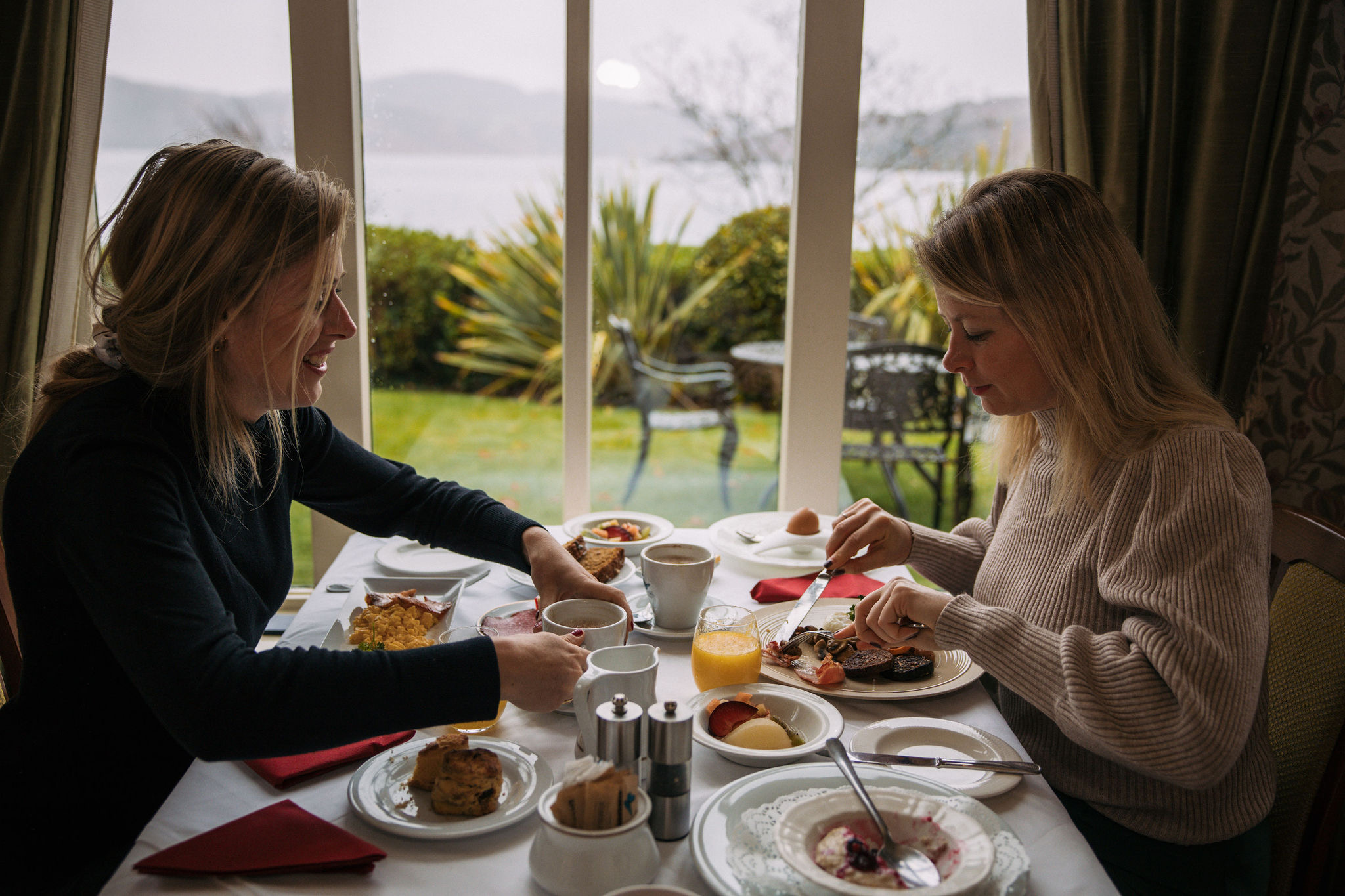 Twee mensen zitten aan een eettafel bij een groot raam met uitzicht op een tuin en water, wat doet denken aan serene rondreis Ierland ochtenden. De tafel is gedekt met ontbijtproducten zoals sinaasappelsap, muffins en jam. Beiden zijn in gesprek en genieten van hun maaltijd in de warme sfeer. | Echt Ierland Vakanties