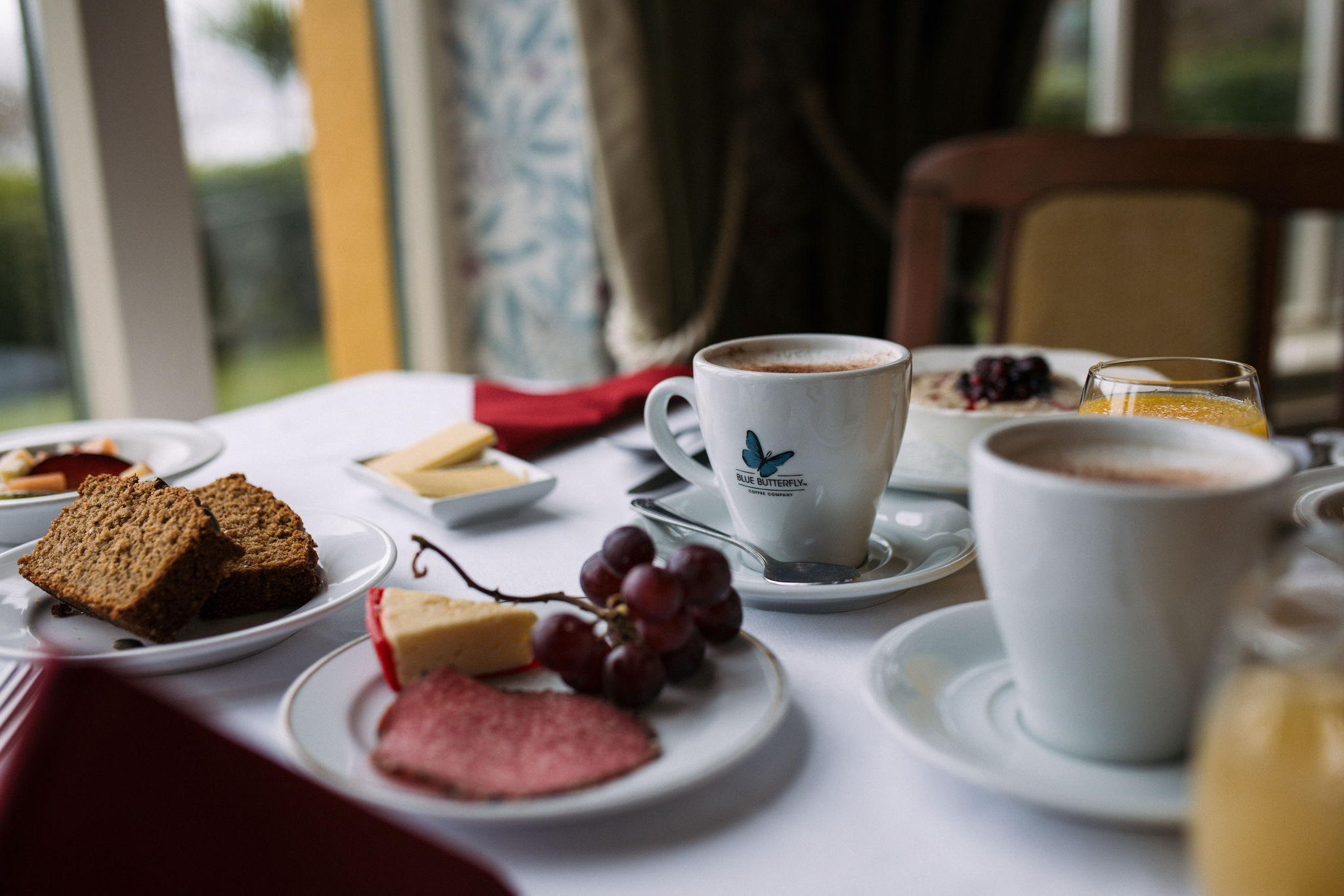 Een ontbijttafel met twee kopjes koffie, borden met gesneden bruin brood, kaas, salami en een klein trosje druiven. Een glas sinaasappelsap op de achtergrond. Het is een gezellige start van uw dag voordat u de schoonheid van Ierland vakanties ontdekt onder het zachte natuurlijke licht van een nabijgelegen raam. | Echt Ierland Vakanties