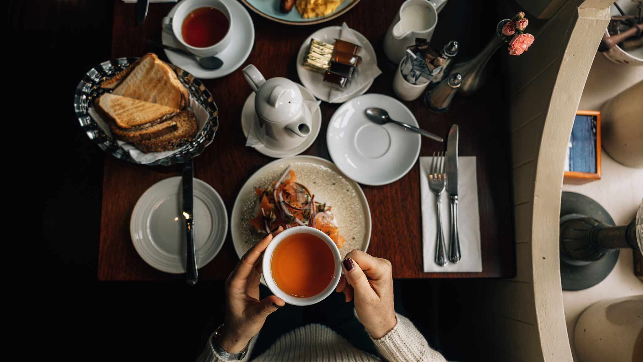 Een persoon houdt een theekopje gevuld met oranje thee boven een houten tafel, wat doet denken aan gezellige vakanties in Ierland. Omringende items omvatten een theepot, toast in een mandje, gerookte zalm met groenten, kleine stukken cake, rode thee en keukengerei, wat een uitnodigende sfeer creëert. | Echt Ierland Vakanties