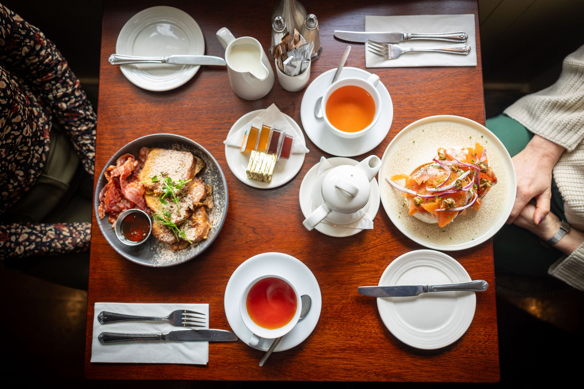 Een houten tafel gedekt voor brunch roept de warmte op van een Ierse vakantie. Borden tonen toast met spek en worstjes, pannenkoeken met fruit en een kleine selectie taart. Kopjes thee en een theepot nodigen u uit om te genieten van momenten die doen denken aan vakanties in Ierland. | Echt Ierland Vakanties