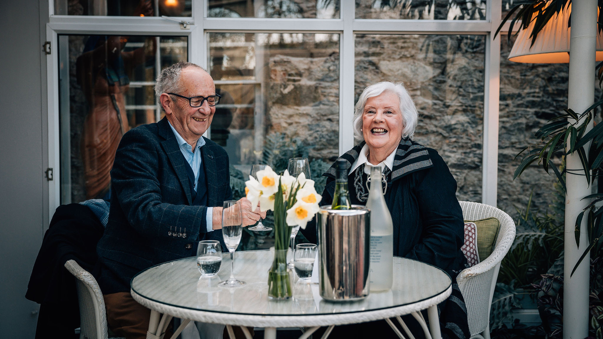 Een ouder echtpaar zit aan een ronde tafel in een zonnige kamer met grote ramen en mijmert over hun onvergetelijke rondreis Ierland. Ze glimlachen hartelijk, narcissen in een vaas naast glazen water en een gekoelde fles in een ijsemmer. Een lamp en planten accentueren de gezellige achtergrond. | Echt Ierland Vakanties