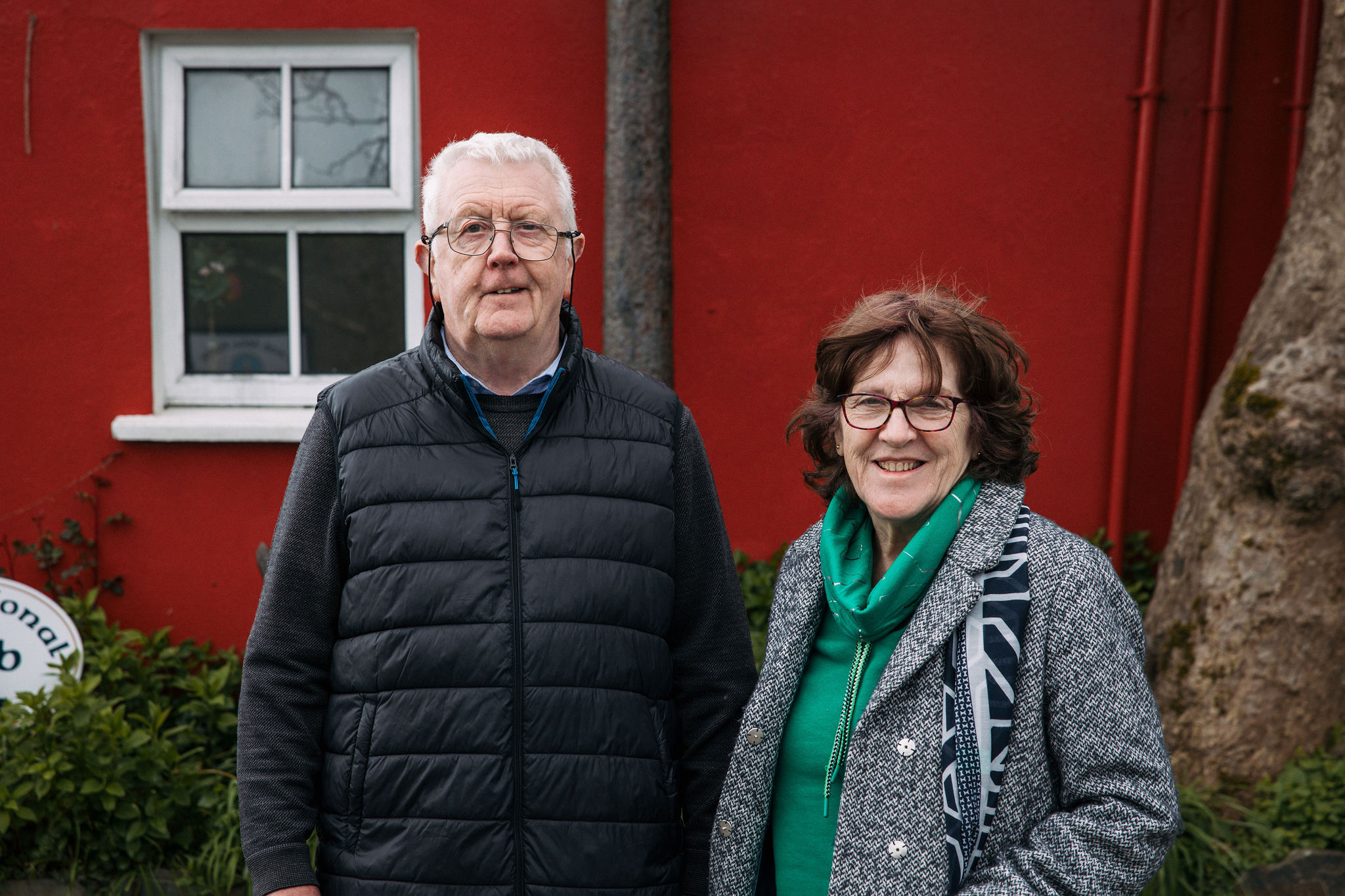 Een oudere man en vrouw staan lachend voor een rood gebouw met een klein raam, wat doet denken aan hun recente rondreis Ierland. De man draagt een bril, een zwart gewatteerd vest en een blauw shirt. De vrouw draagt een bril, een groene sjaal en een grijze jas. Een boomstam en gedeeltelijk zichtbaar wit bord zijn op de achtergrond te zien. | Echt Ierland Vakanties