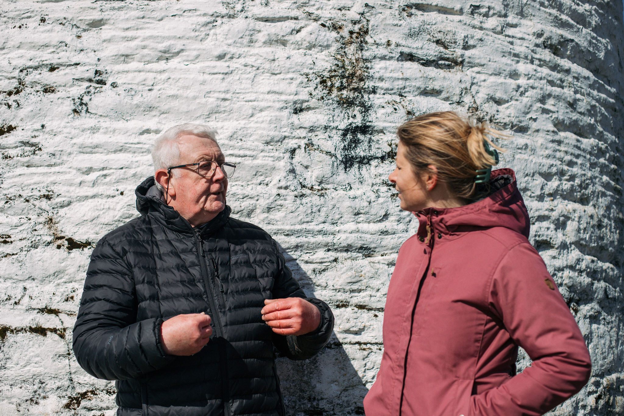 Een oudere man in een zwart jasje en een bril bespreekt geanimeerd hun laatste rondreis Ierland met een vrouw in een rood jasje. Ze staan buiten tegen een getextureerde witte bakstenen muur, badend in zonlicht, tegenover elkaar en in gesprek. | Echt Ierland Vakanties