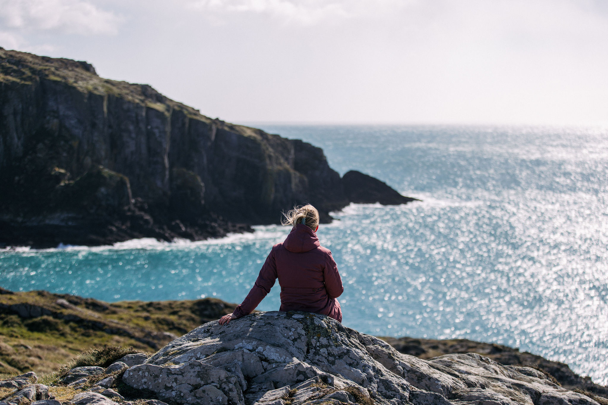 Een persoon met blond haar in een rood jasje zit op een grote rots, met uitzicht op de oceaan tijdens hun vakantie in Ierland. Ze kijken uit over een blauwe zee met zachte golven en ruige kliffen op de achtergrond. De lucht is gedeeltelijk bewolkt, waardoor er een zacht licht over dit schilderachtige landschap werpt, perfect voor een vakantie in Ierland. | Echt Ierland Vakanties