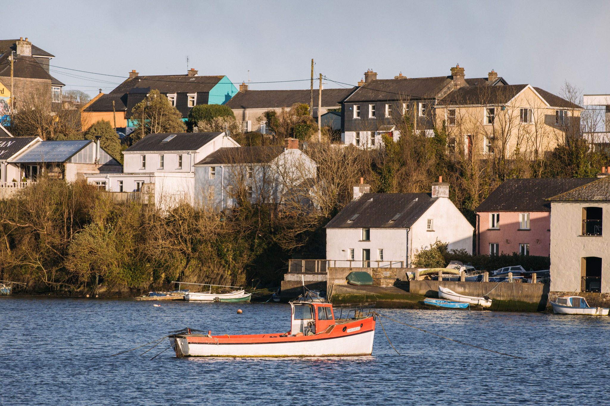 Een kleine rood-witte vissersboot ligt voor anker op het kalme blauwe water vlak bij de kust. Op de achtergrond staan verschillende huizen in gedempte kleuren, genesteld tussen bladloze bomen en struiken op een zacht glooiende heuvel onder een heldere hemel. Een pittoresk tafereel dat perfect is voor elke rondreis Ierland. | Echt Ierland Vakanties