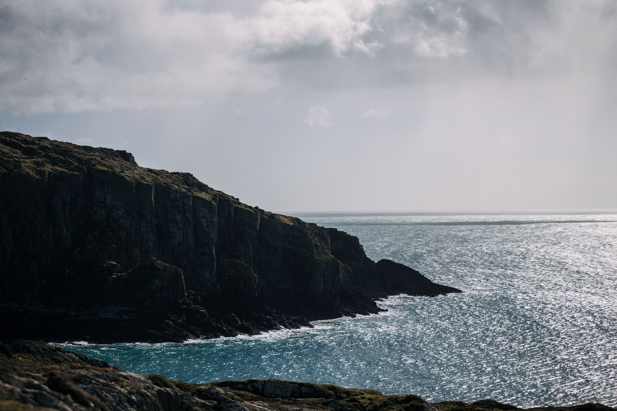 Rotsachtige kustkliffen in Ierland kijken uit over een uitgestrekt, glinsterend waterlichaam onder een bewolkte lucht. De ruige klif is versierd met stukken gras en mos. Zonlicht filtert door de wolken en creëert heldere reflecties op het wateroppervlak, wat een serene maar dramatische sfeer biedt die perfect is voor een vakantie in Ierland. | Echt Ierland Vakanties