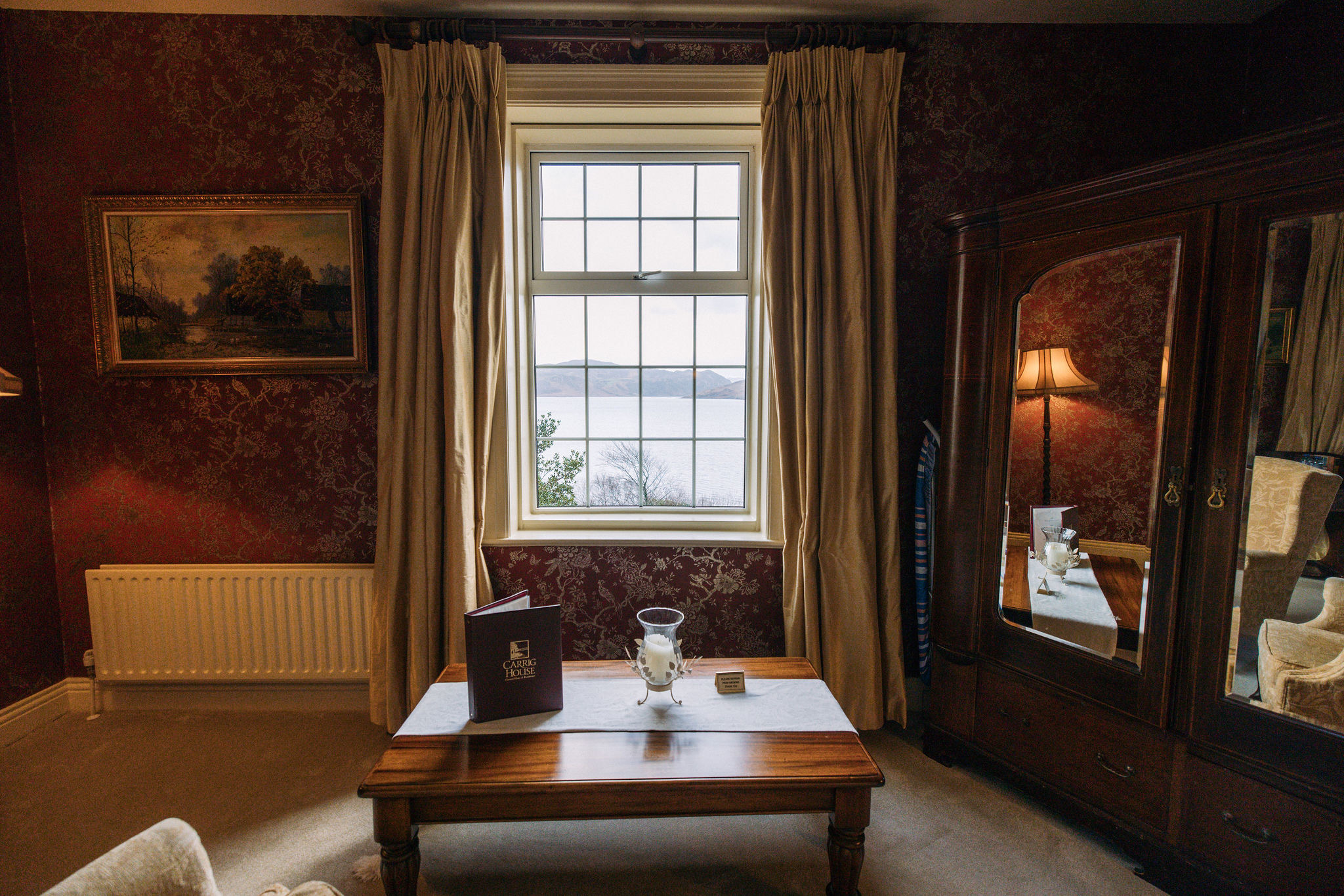 Een gezellige kamer met een centraal raam met uitzicht op het meer, perfect voor degenen die dromen van een vakantie in Ierland. De muren zijn voorzien van donker bloemenbehang en een landschapsschilderij. Een houten tafel met een menu en een kaars, terwijl een spiegelkast en lampenstandaard aan de rechterkant staan. Crèmekleurige gordijnen en een radiator maken de setting compleet. | Echt Ierland Vakanties