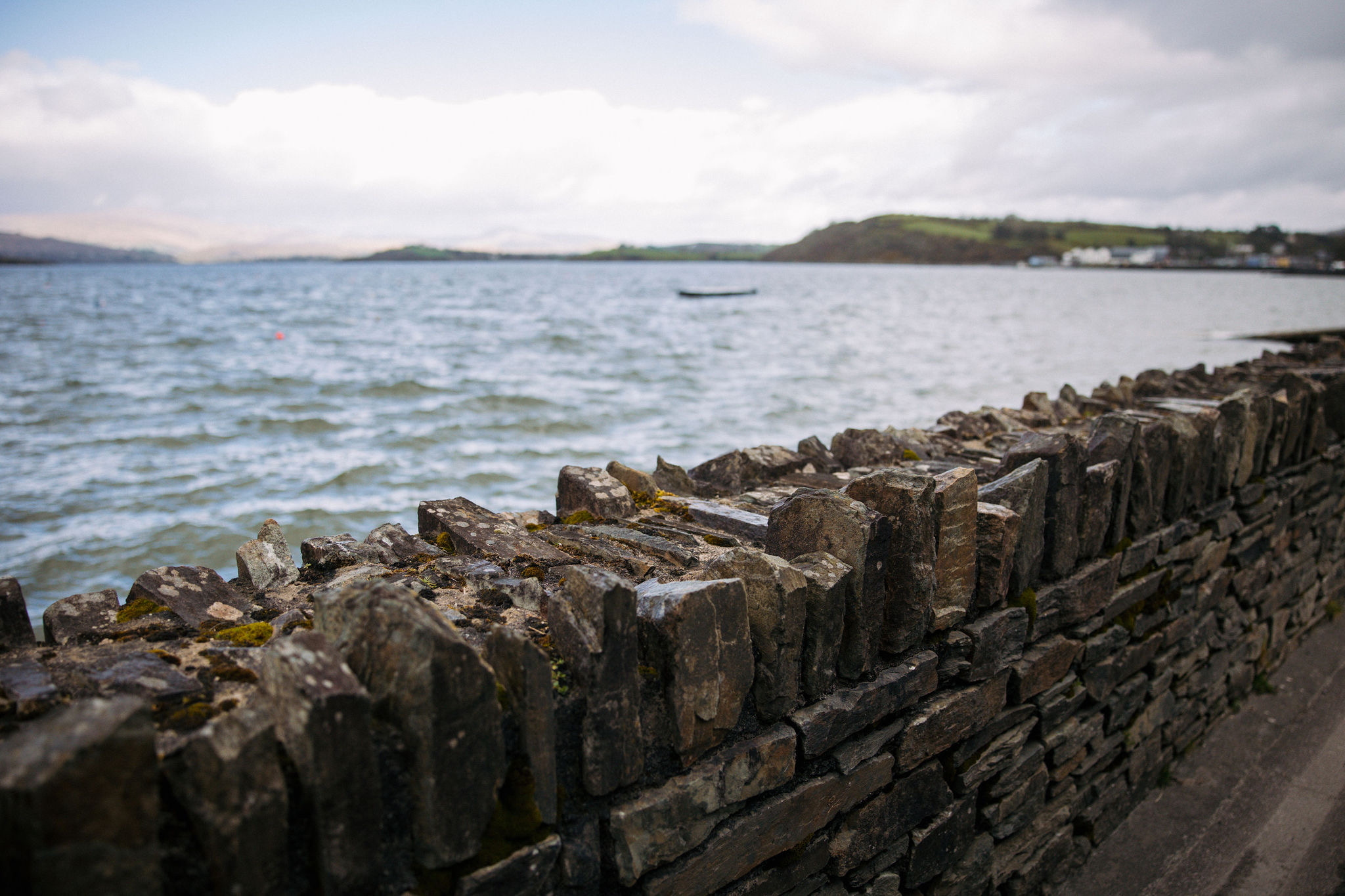 Een stenen muur omlijnt de rand van een waterkant, waar de choppy golven van een uitgestrekt waterlichaam de kust ontmoeten - een glimp van wat u te wachten staat tijdens een onvergetelijke vakantie in Ierland. In de verte vaart een kleine boot onder een bewolkte lucht, met op de achtergrond heuvelachtige landschappen en schaarse gebouwen langs de kustlijn. | Echt Ierland Vakanties