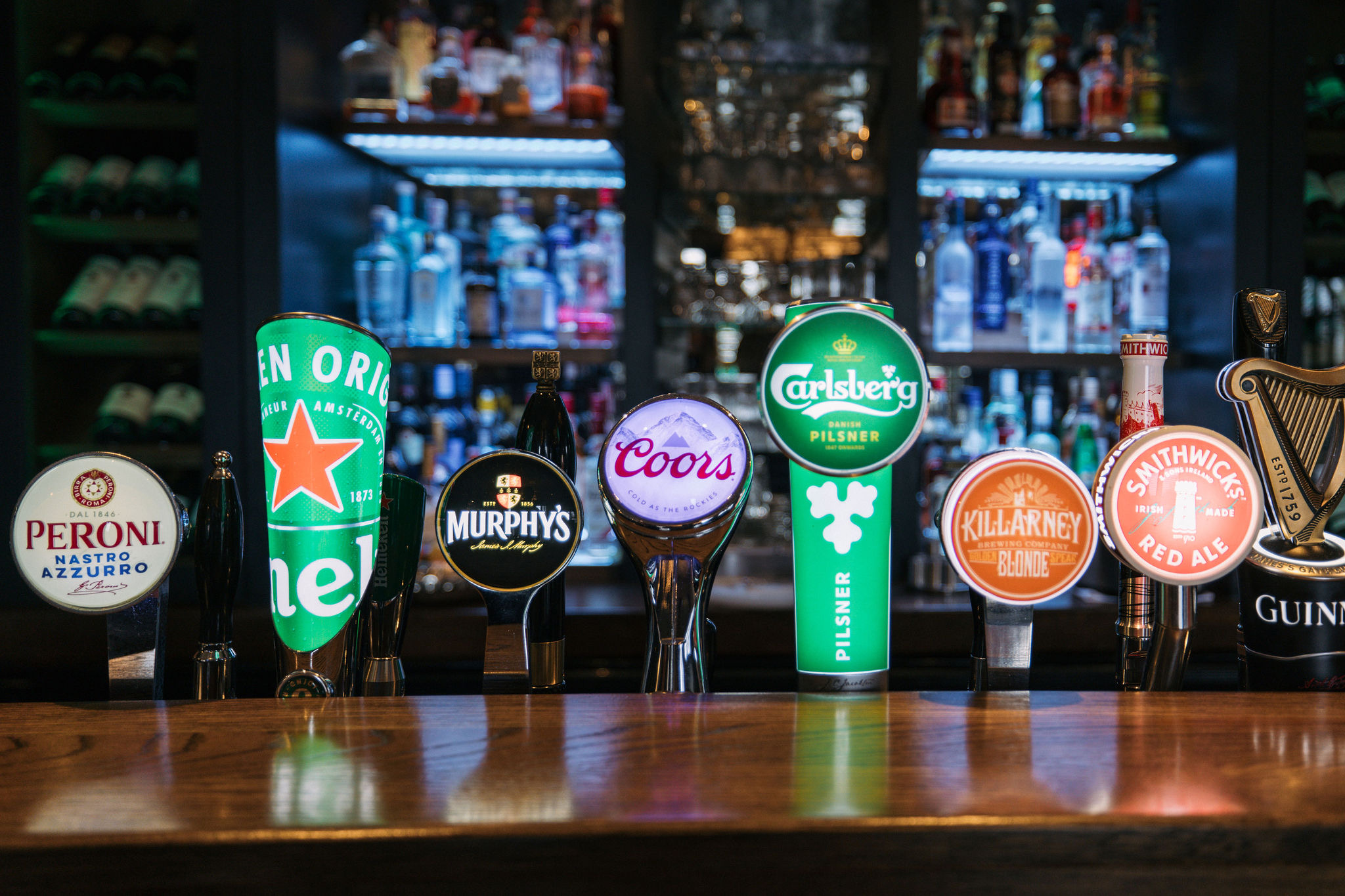 Een schemerige bar met verschillende biertaps op een houten toonbank, die een echte 'Ierland vakantie'-smaak bieden. Merken als Peroni, Heineken en Guinness springen in het oog. Planken op de achtergrond tonen flessen sterke drank, wat bijdraagt aan de warme, uitnodigende sfeer die doet denken aan een rondreis Ierland. | Echt Ierland Vakanties