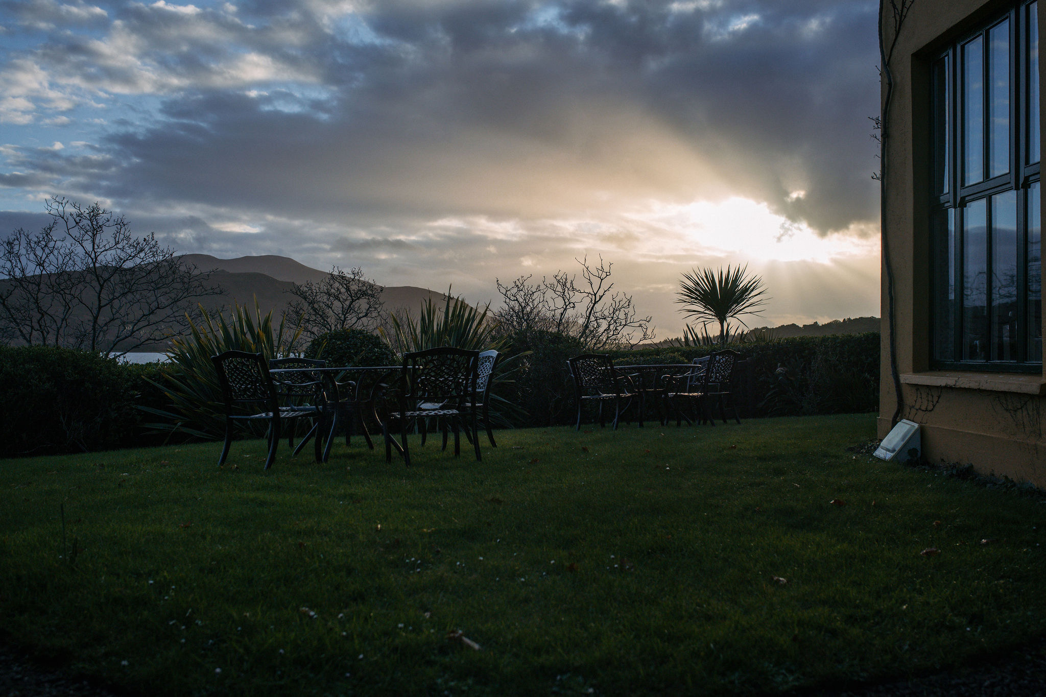 Buitenscène bij zonsondergang met metalen stoelen en tafels op een grasveld roept de charme van vakanties in Ierland op. De zonnestralen doorboren de wolken boven verre heuvels en creëren een dramatische lucht. De hoek van een gebouw met grote ramen bevindt zich aan de rechterkant, terwijl bomen en struiken deze serene Ierse sfeer versterken. | Echt Ierland Vakanties