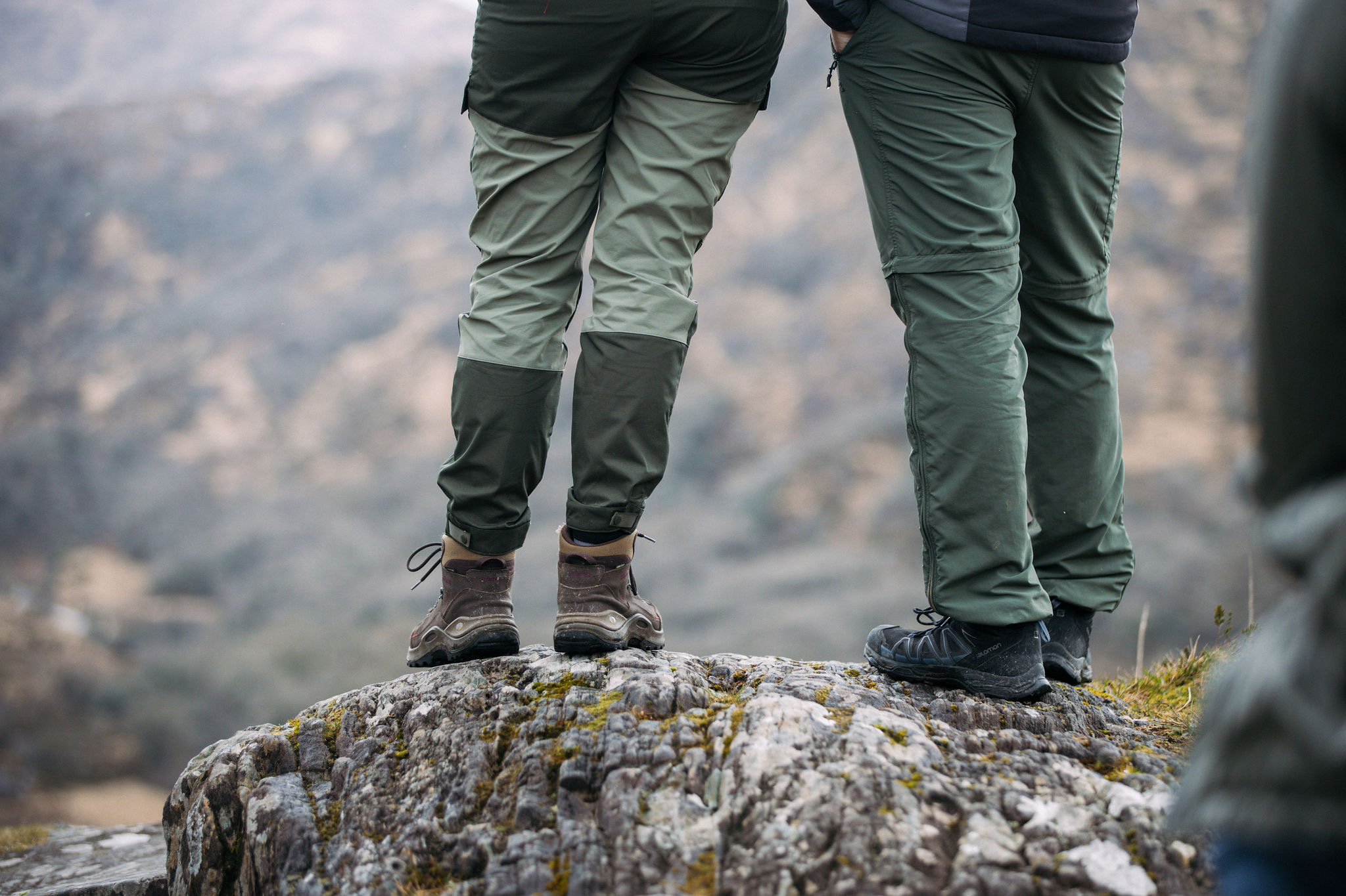 Twee mensen staan op een rotspunt, gekleed in een outdoorbroek en wandelschoenen, en vangen de essentie van een avontuurlijke vakantie in Ierland. De wazige heuvels op de achtergrond suggereren een betoverende natuuromgeving, met weelderige groene broeken en stevige laarzen die de aandacht stelen. | Echt Ierland Vakanties