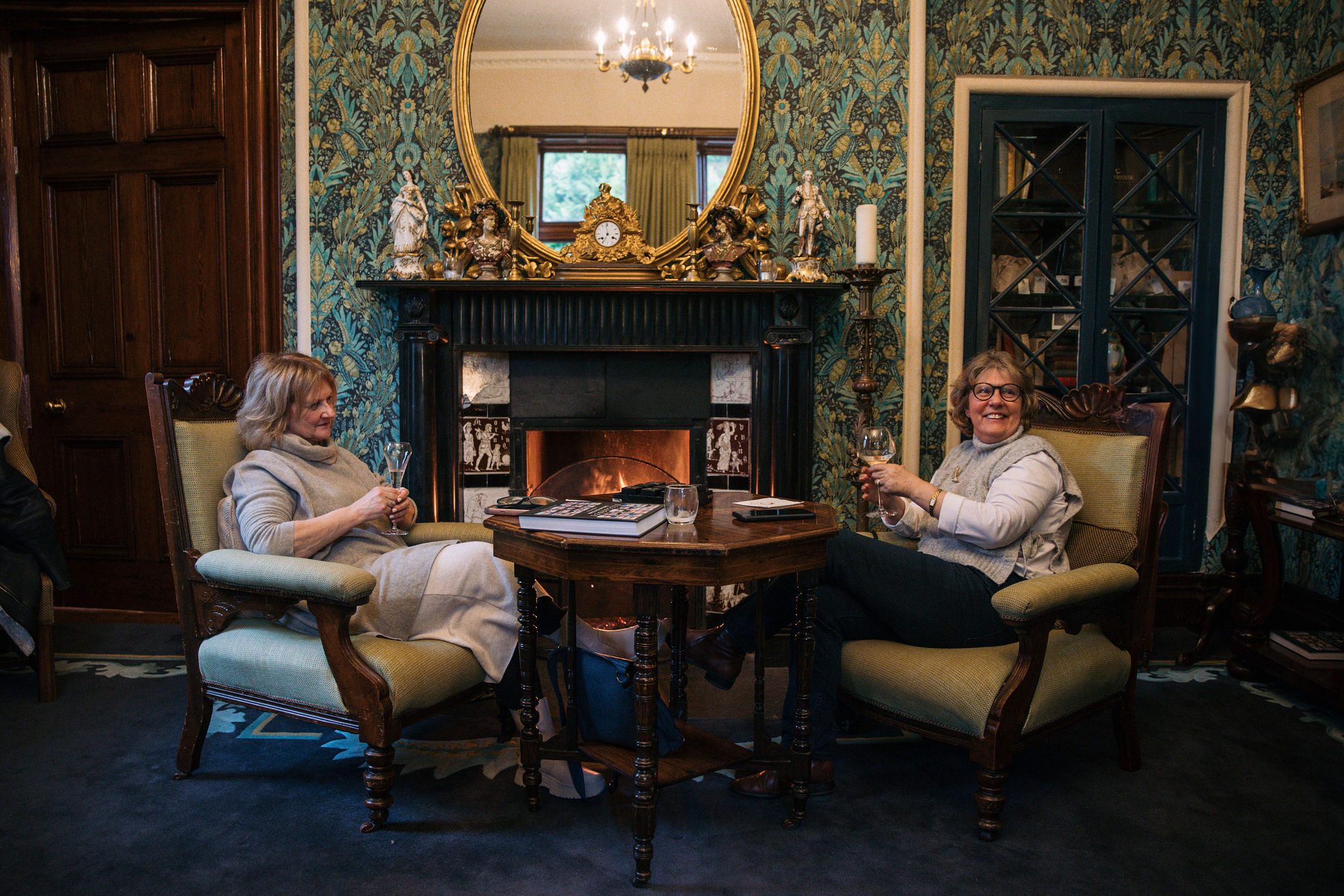 Twee vrouwen zitten in fauteuils bij een brandende open haard in een gezellige, rijkelijk versierde kamer, die doet denken aan een uitnodigend huisje dat ze vonden tijdens hun rondreis door Ierland. Ze glimlachen terwijl ze kopjes vasthouden. Bloemenbehang, een grote spiegel boven de open haard en planken met decoratieve items maken het tafereel compleet. | Echt Ierland Vakanties