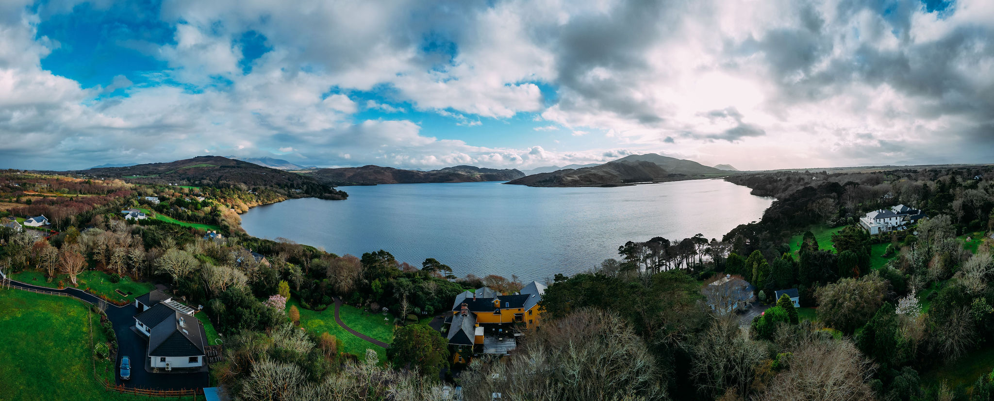 Een panoramisch uitzicht op een sereen meer in Ierland, omgeven door weelderig groen en verre heuvels onder een gedeeltelijk bewolkte lucht. Dichtbij de kust steken verschillende huizen met donkere en oranje daken door de bomen heen. Het water weerspiegelt de lucht, wat een rustig gevoel toevoegt aan de ideale scène voor een rondreis Ierland. | Echt Ierland Vakanties