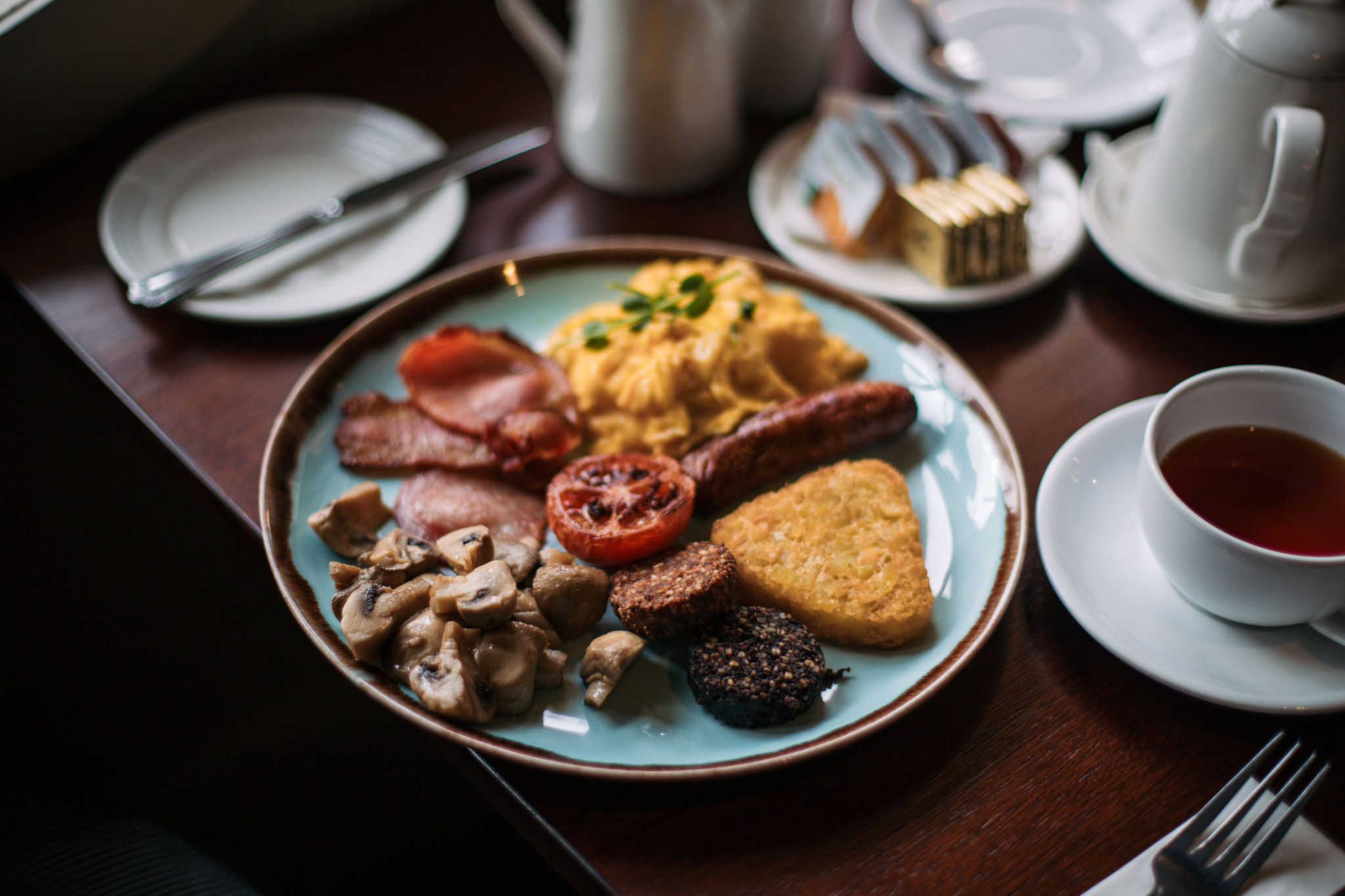 Een volledig Engels ontbijt op een bord bestaat uit knapperig spek, roerei, worst, gegrilde tomatenschijfjes, hash brown, champignons en bloedworst. Het past perfect bij herinneringen aan vakantie in Ierland terwijl u thee drinkt van een schoteltje naast uw houten tafel met kleine bordjes en bestek. | Echt Ierland Vakanties