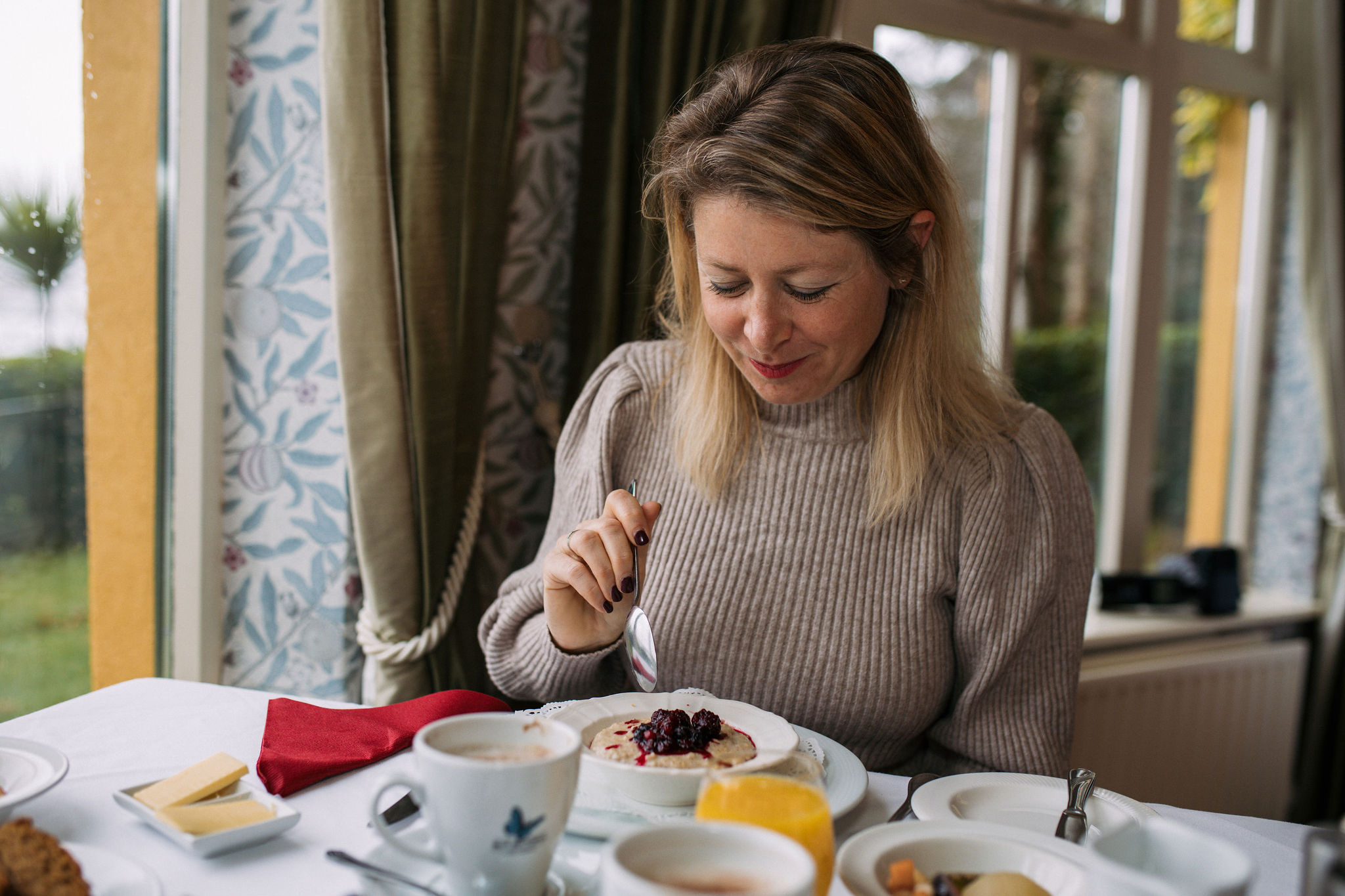 Een vrouw met blond haar, gekleed in een grijze trui, zit aan een tafeltje bij het raam en geniet van haar dessert. Met een glimlach en een lepel in haar hand geniet ze van de lekkernijen die haar aan haar vakantie in Ierland doen denken. De achtergrond toont gordijnen met patronen en een prachtig uitzicht op de tuin. | Echt Ierland Vakanties