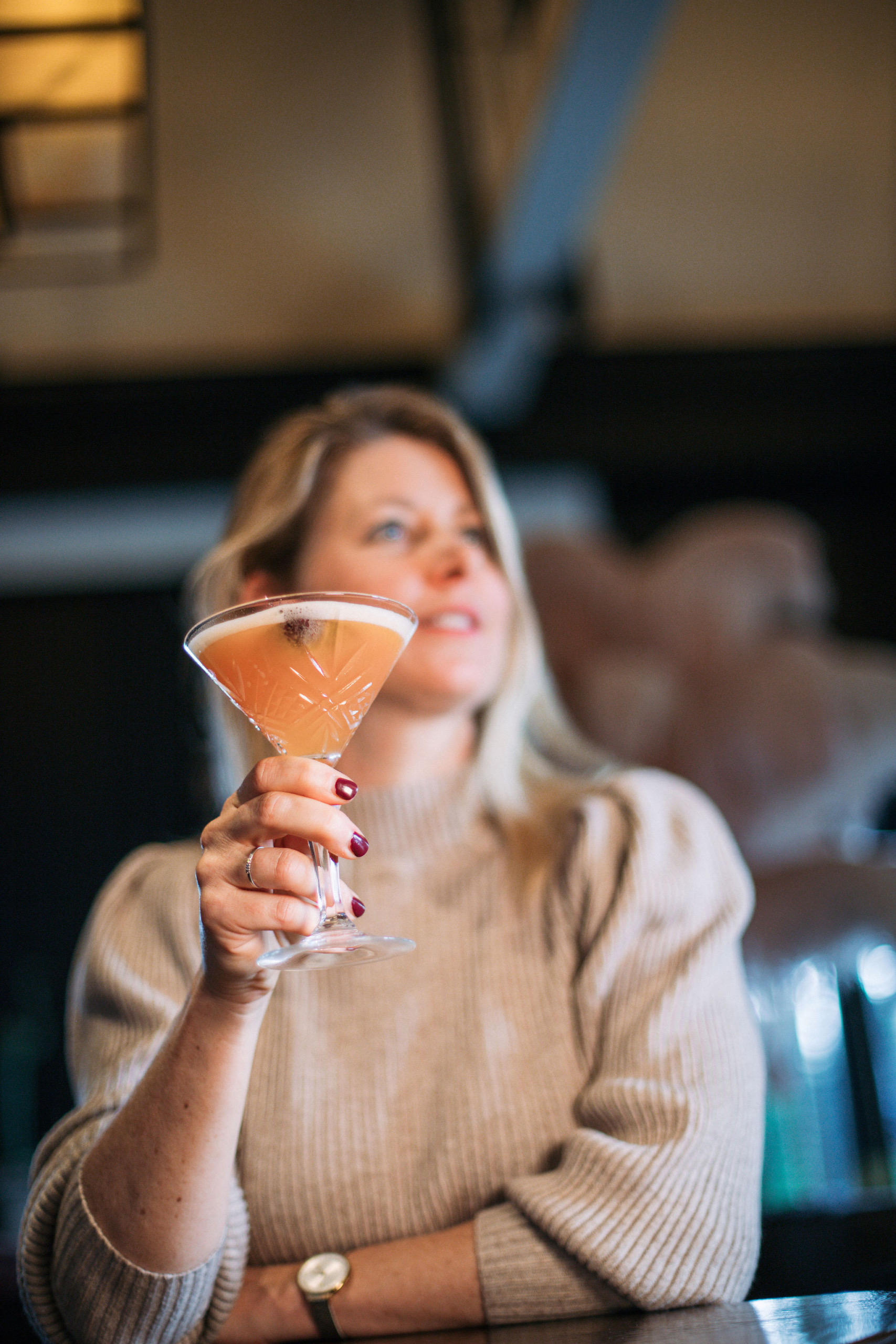 Een vrouw in een beige trui houdt een martiniglas vast met een roze cocktail, starend in de verte alsof ze droomt van een vakantie in Ierland. Haar lichte haar complimenteert haar horloge, terwijl de zacht wazige achtergrond en warme verlichting wijzen op een binnenomgeving, misschien een gezellige bar of restaurant. | Echt Ierland Vakanties
