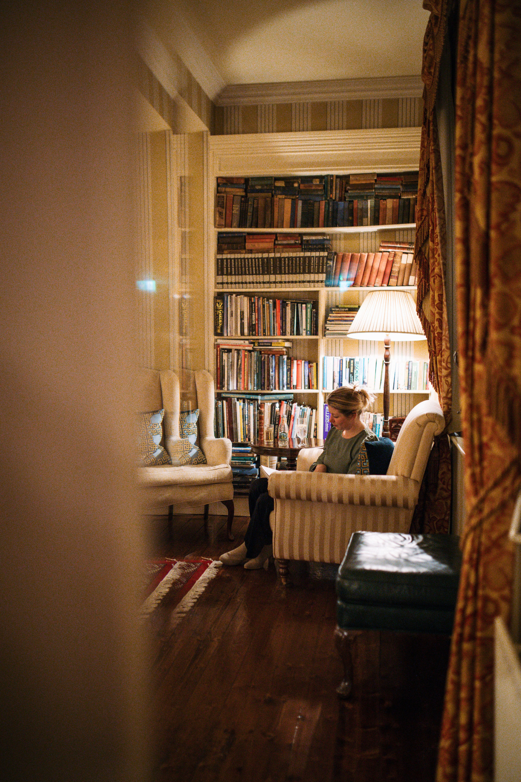 Een persoon ontspant in een gezellige, goed verlichte kamer die doet denken aan een vakantie in Ierland. Omringd door boekenplanken vol verhalen, zitten ze in een fauteuil naast een lamp, waarvan de gloed warmte toevoegt aan de gepatroneerde gordijnen en houten vloer. Een andere gewatteerde fauteuil wacht in de buurt voor dromerige ontsnappingsplannen. | Echt Ierland Vakanties