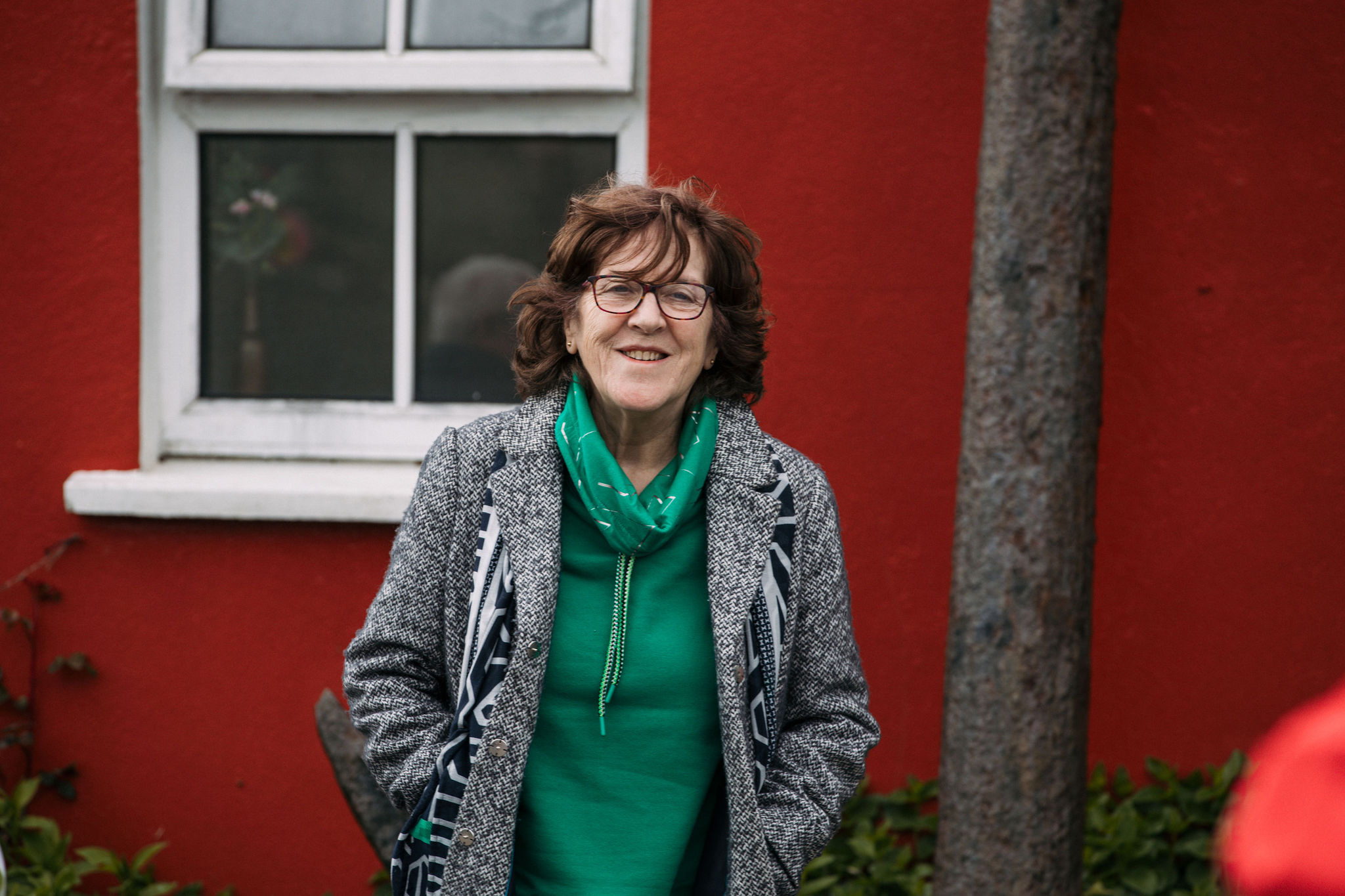 Een vrouw met een bril, een grijze jas en een groene sjaal over een groen topje, staat lachend voor een felrode muur met een raam, de charme van haar rondreis Ierland vastleggend. Haar handen zitten in haar jaszakken. Een boomstam is naast haar zichtbaar te midden van weelderig groen. | Echt Ierland Vakanties